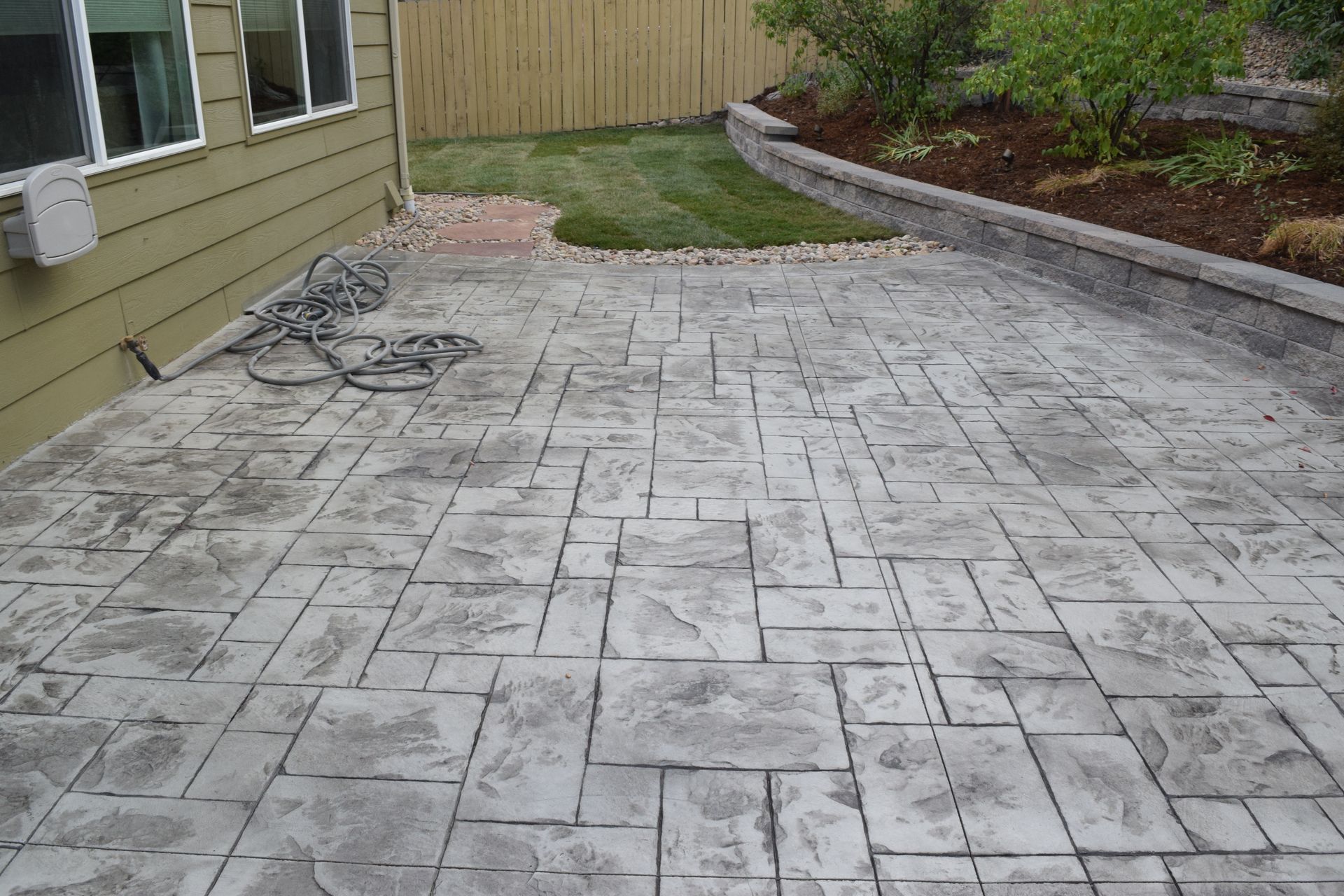 Stamped concrete patio with a gray stone pattern next to a grassy lawn and a retaining wall.