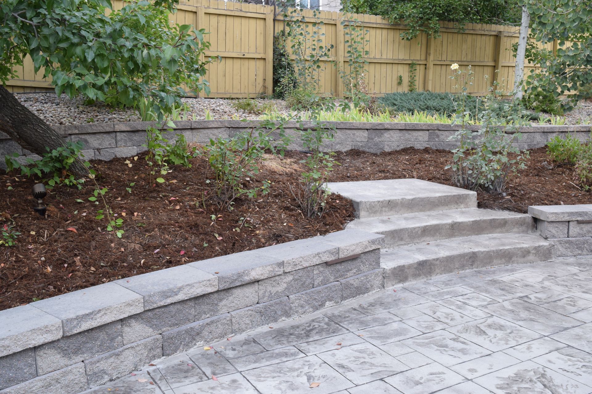 Stone steps and retaining walls in a backyard, leading up to a mulched garden bed and wooden fence.