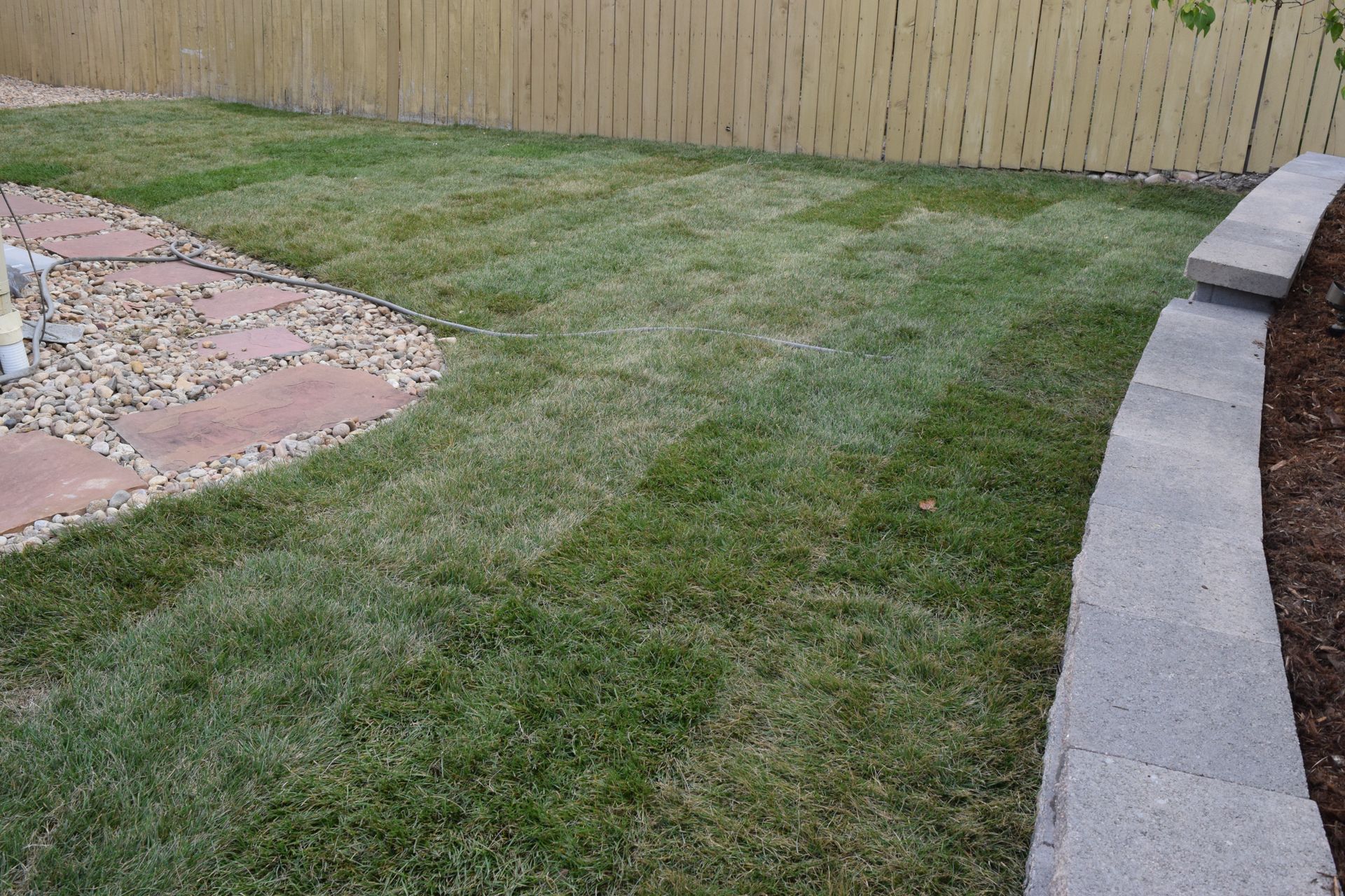 Green lawn with mowing stripes, bordered by a wooden fence, stone pathway, and a retaining wall.