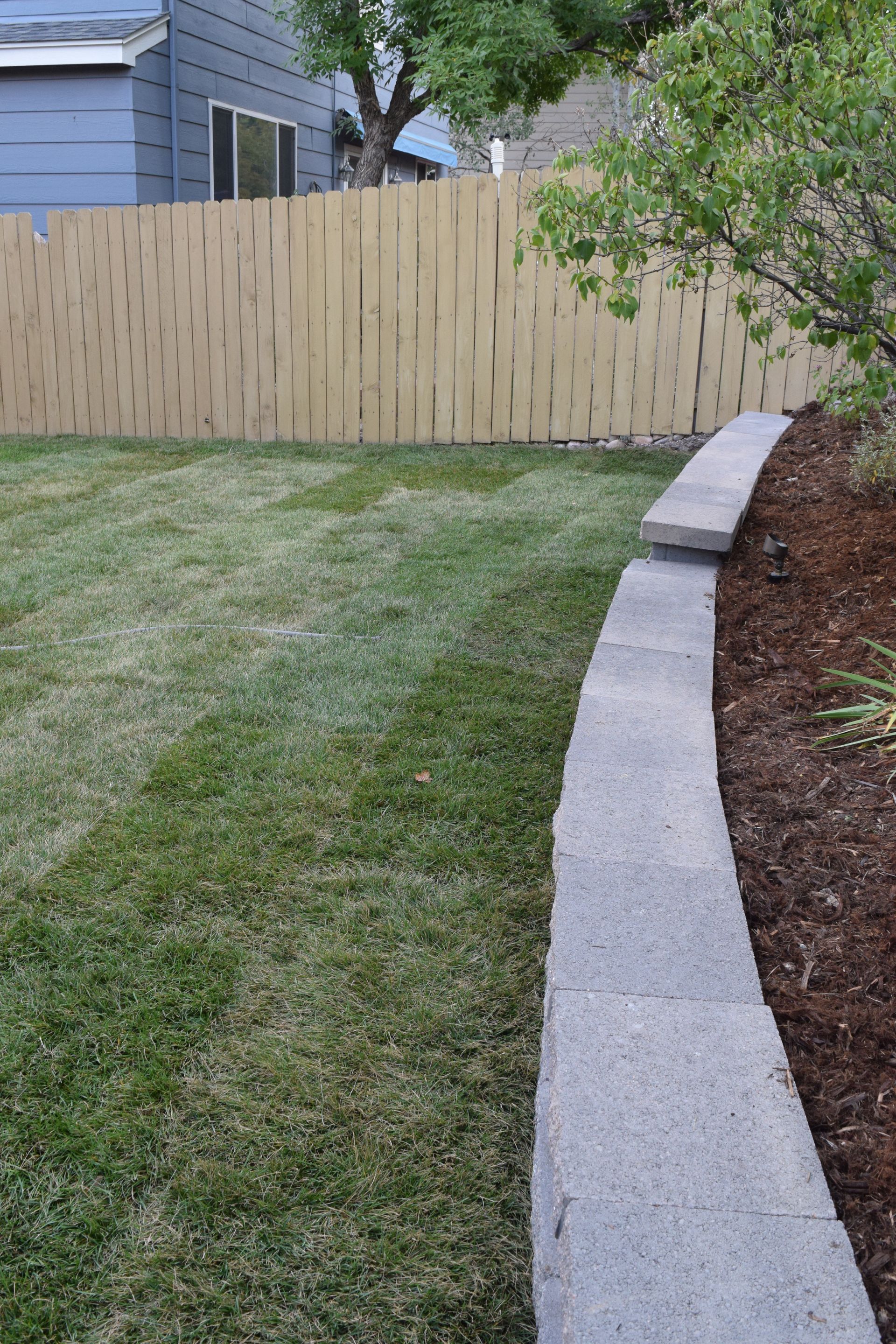Green grass yard with a curved grey stone retaining wall and a wood fence.
