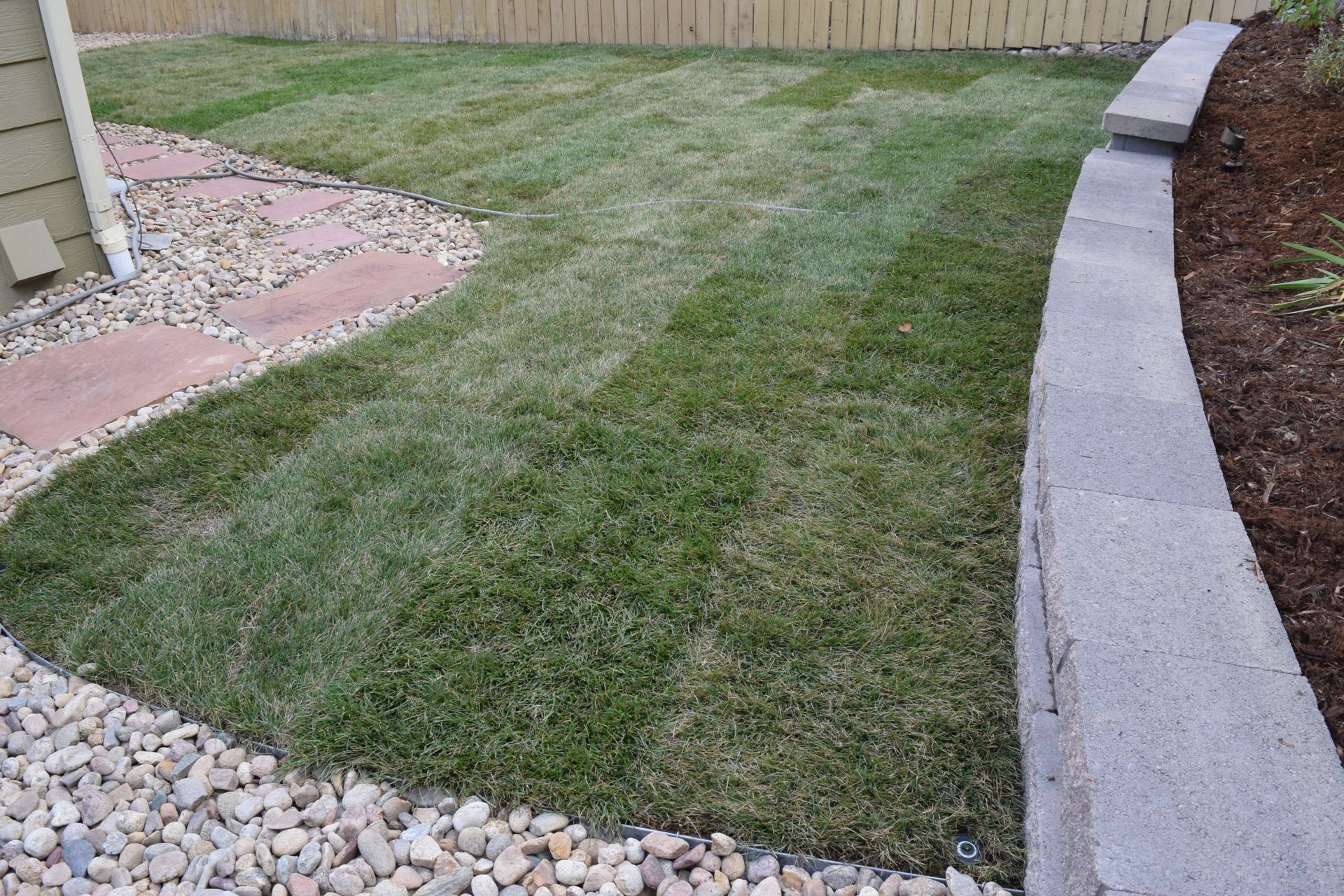 Lawn with distinct mowing patterns, bordered by a stone retaining wall, patio, and gravel.