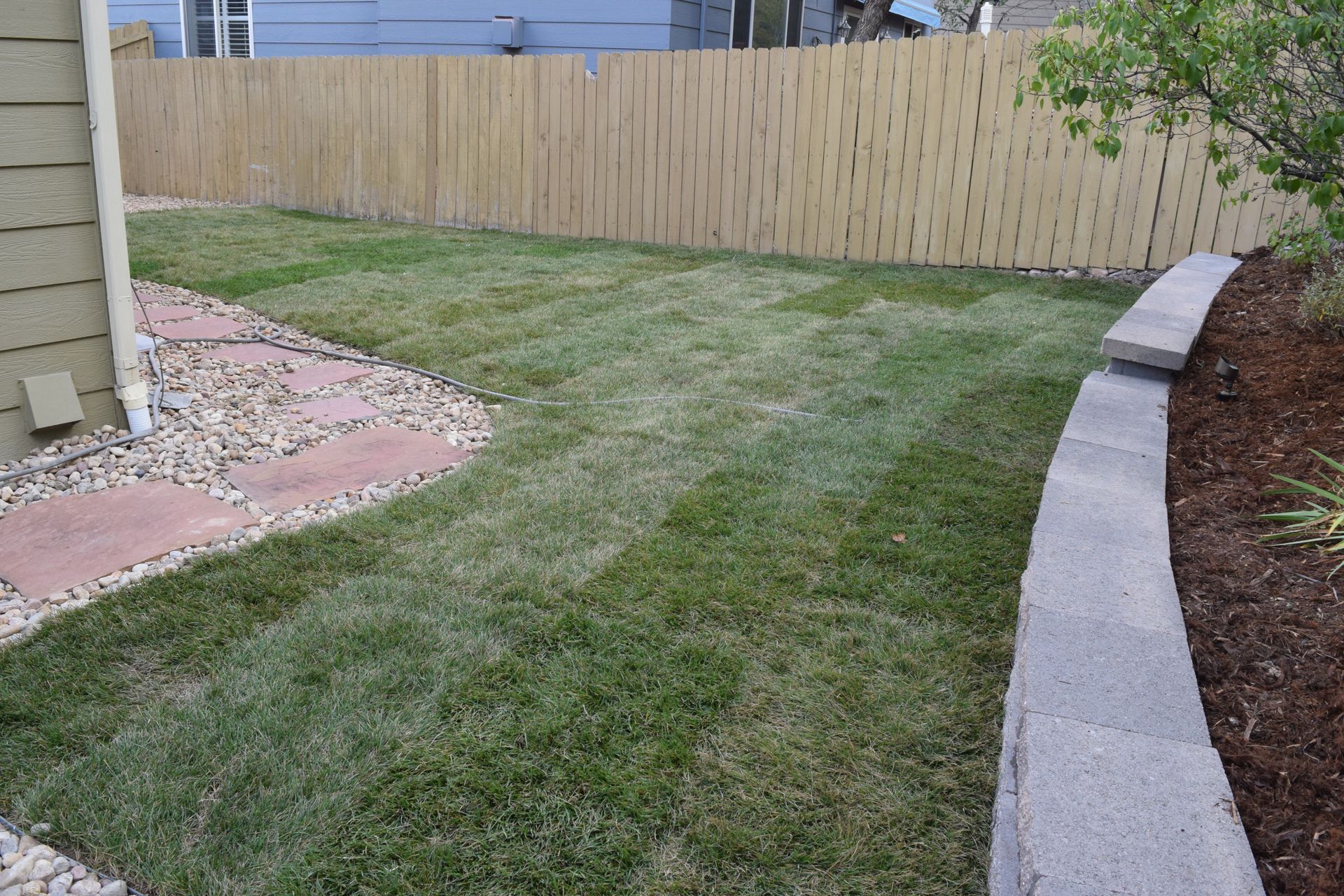 Lawn in a backyard with a stone path, retaining wall, and wooden fence.