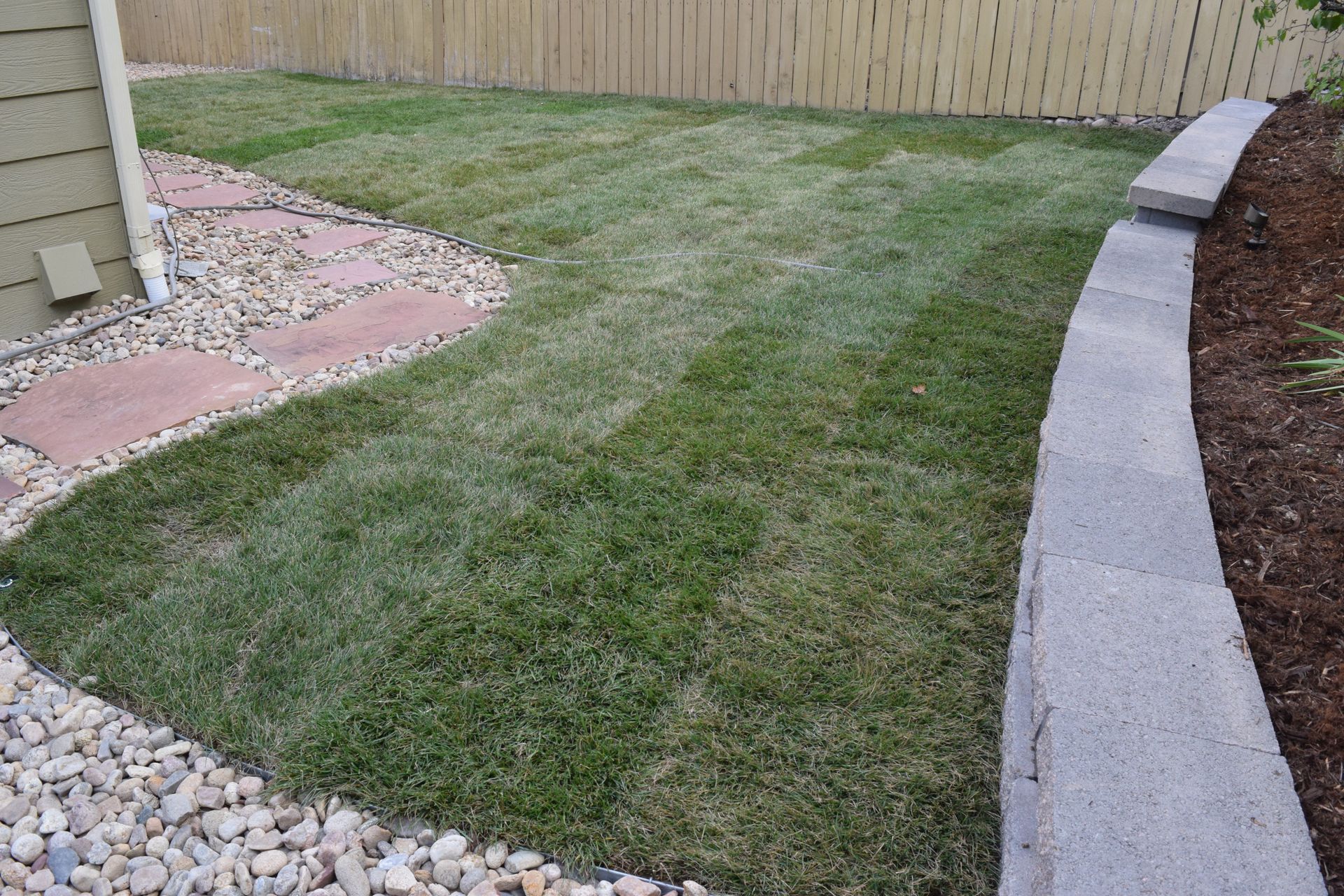 Lawn with fresh cut stripes, bordered by gravel, stone walkway, and a concrete retaining wall, with a wooden fence background.