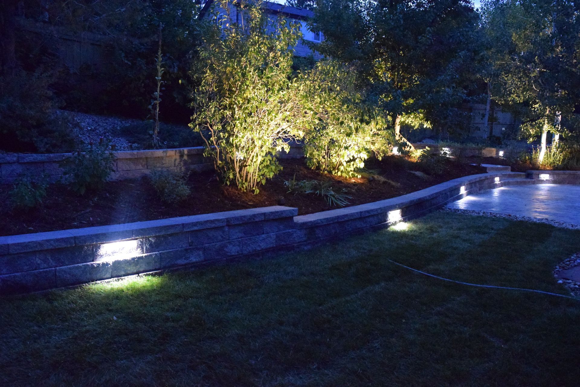 Nighttime outdoor scene with lit trees, a retaining wall with lights, and a grassy area.