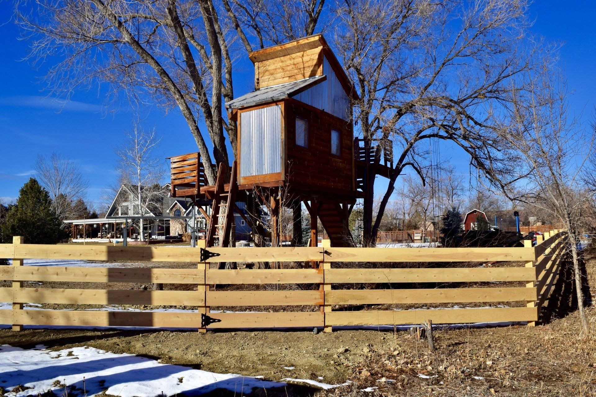 Wooden treehouse in a large tree, behind a wood fence, blue sky, snow patches on ground.