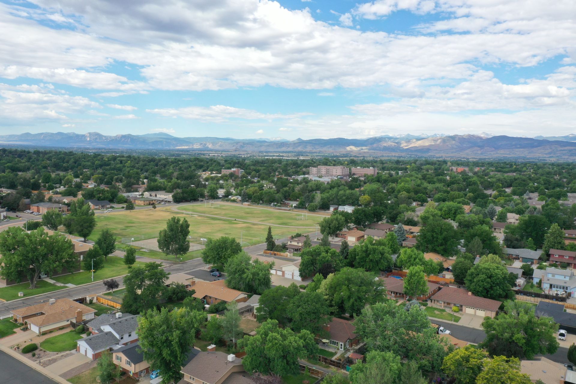 Aerial view of a suburban neighborhood with green trees, houses, and a mountain range in the distance under a cloudy blue sky.