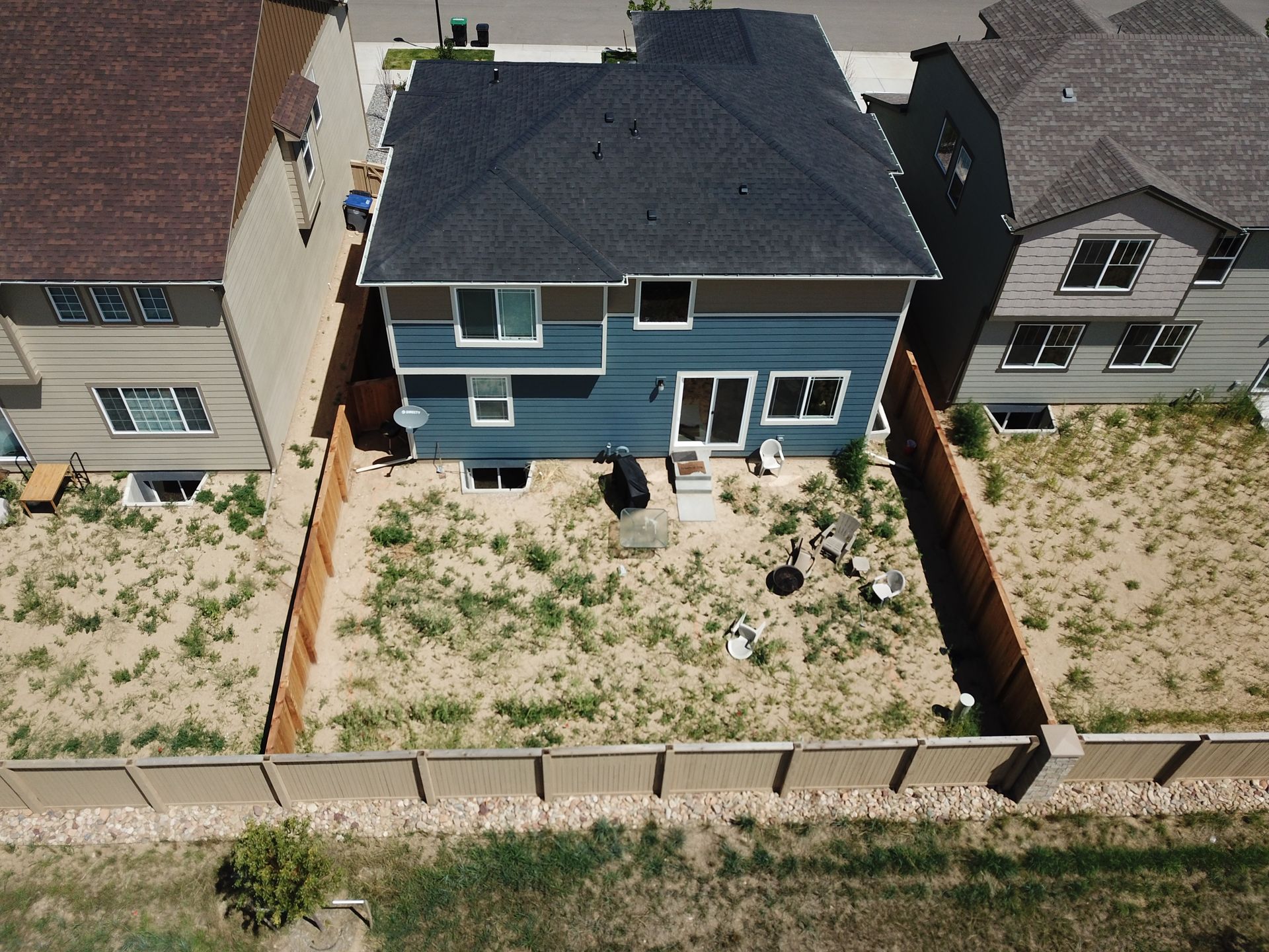 Aerial view of a two-story blue house with a dark roof and a weedy backyard, between two other houses with brown roofs.