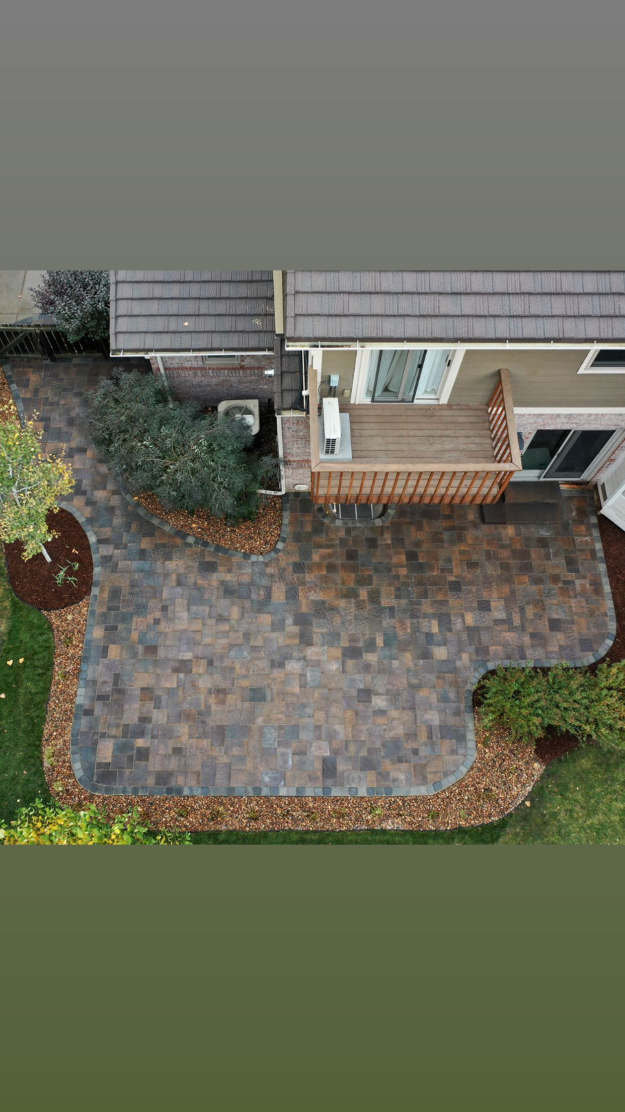 Aerial view: Patio with pavers, small deck, beige house, and surrounding greenery.