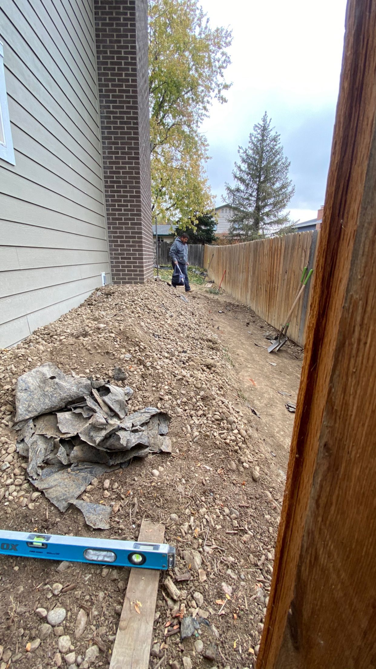 A person works in a dirt and woodchip area next to a building and fence, with a level in the foreground.