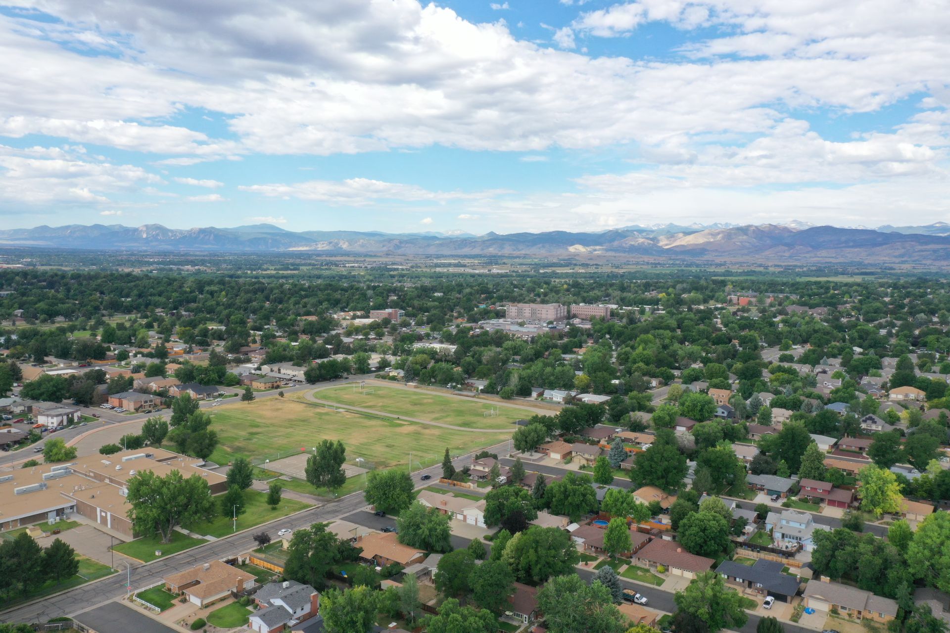 Aerial view of a suburban neighborhood with green trees, buildings, and a cloudy sky in the background with mountains.