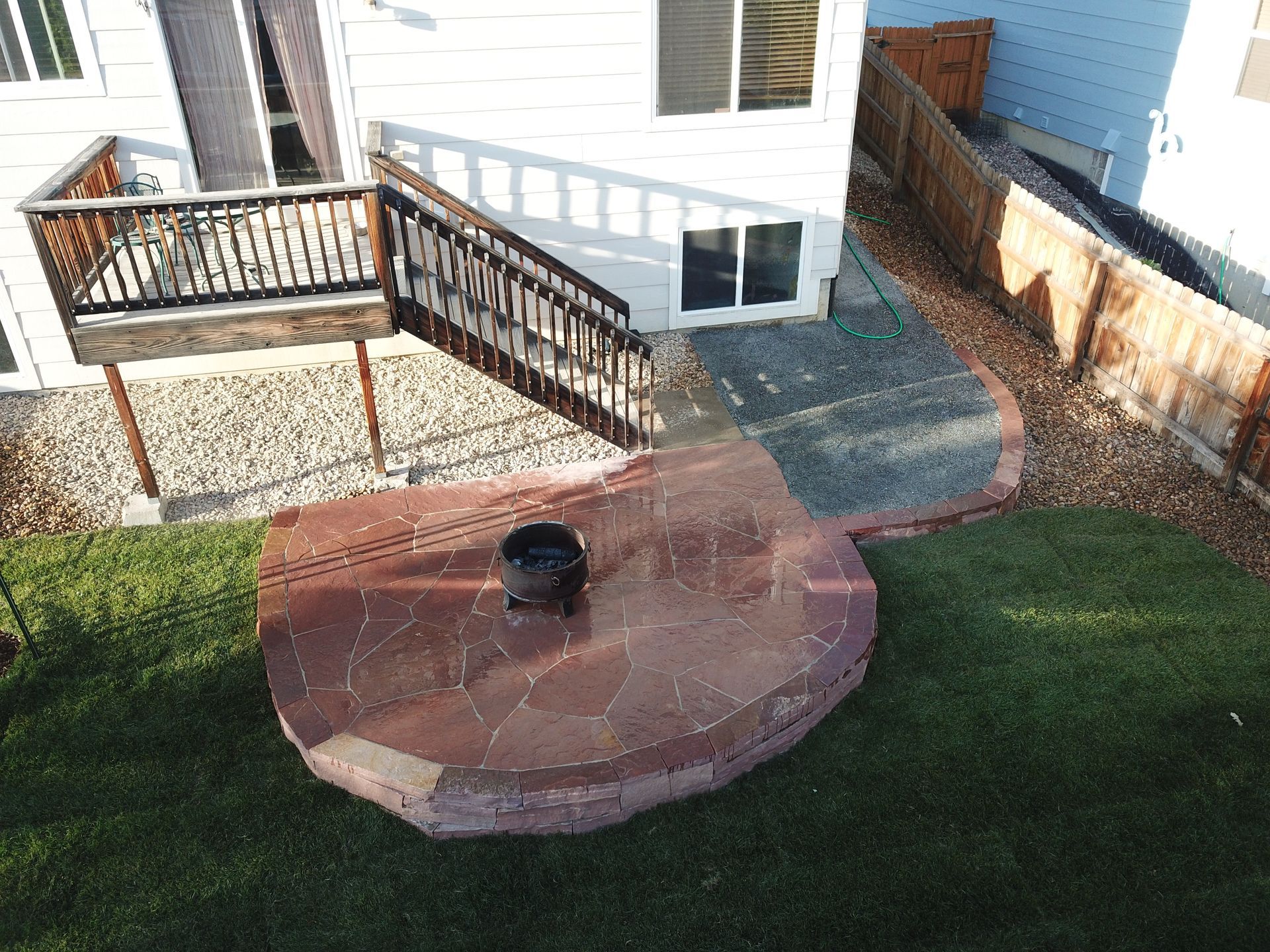 Backyard patio with fire pit, stone base, wooden deck, green grass, and white house.