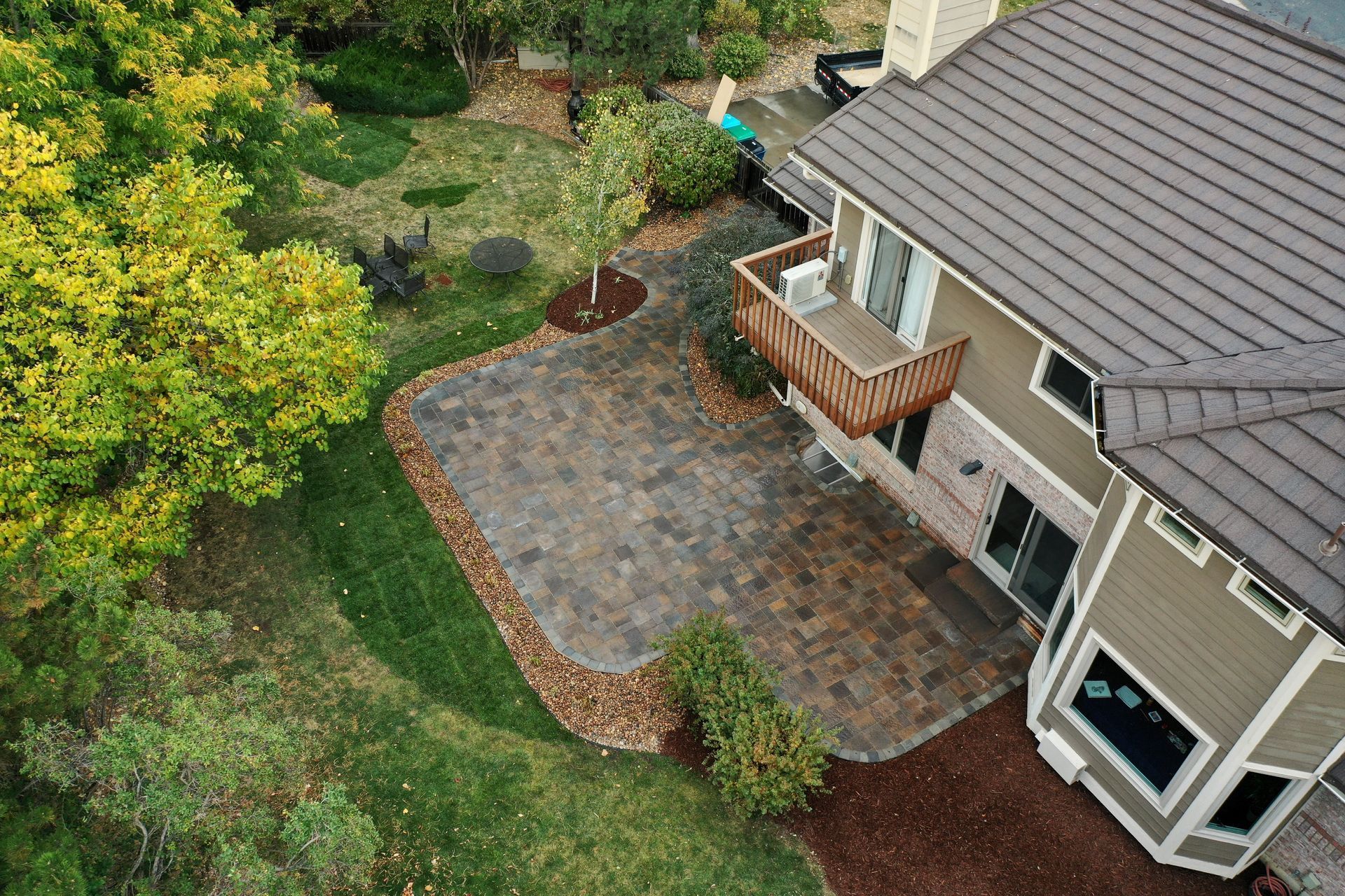Aerial view of a home with a large patio and green lawn. Patio has pavers, house has a brown roof.
