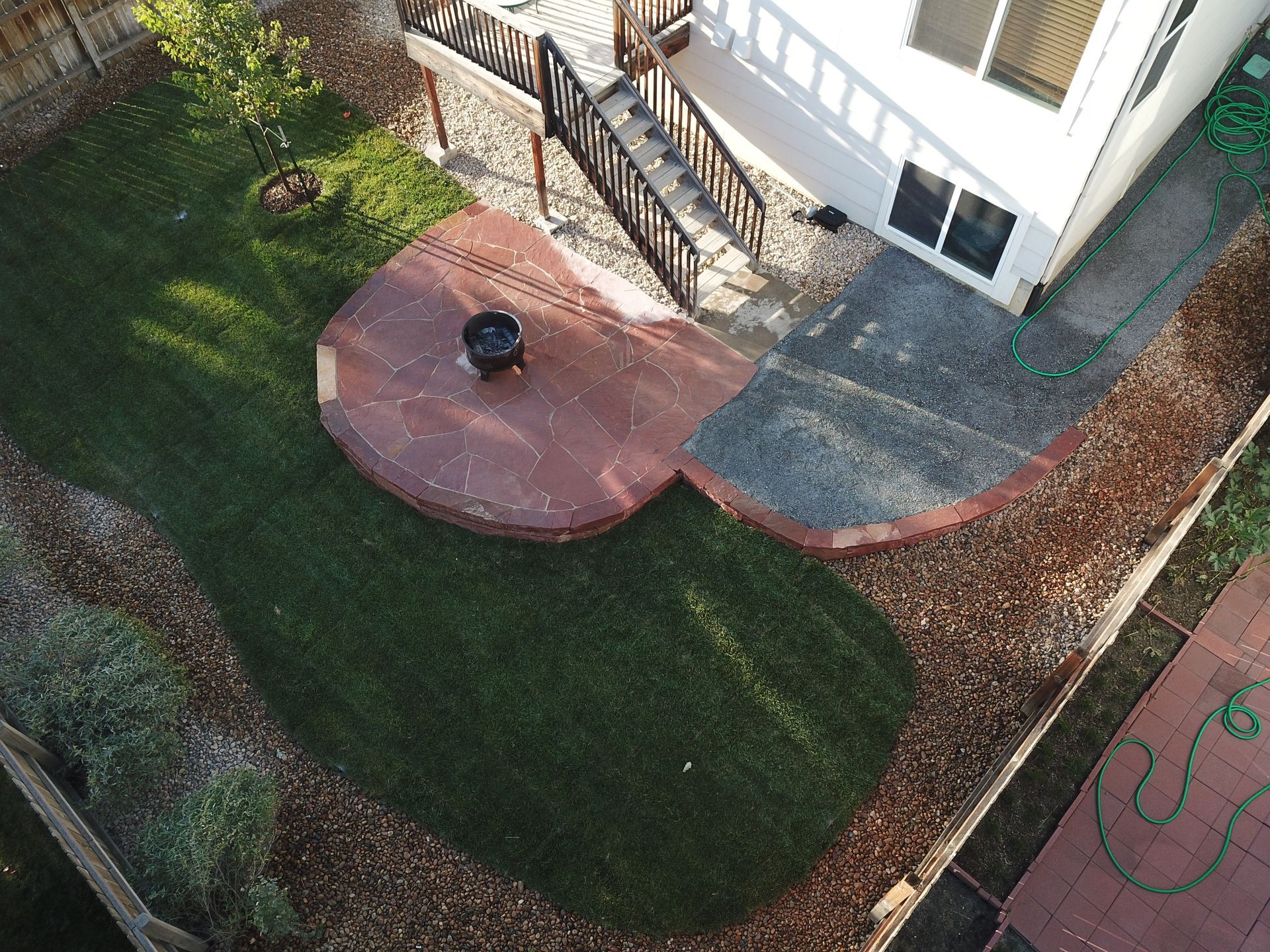 Overhead view of a backyard with a deck, patio, fire pit, and lawn; tan, brown, and green.