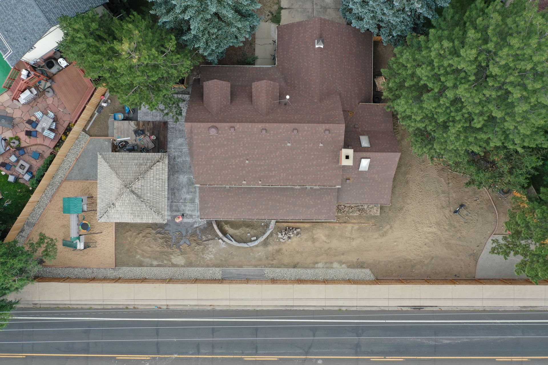 Overhead view of a house with a brown roof, surrounded by trees, a patio, and a road in front.