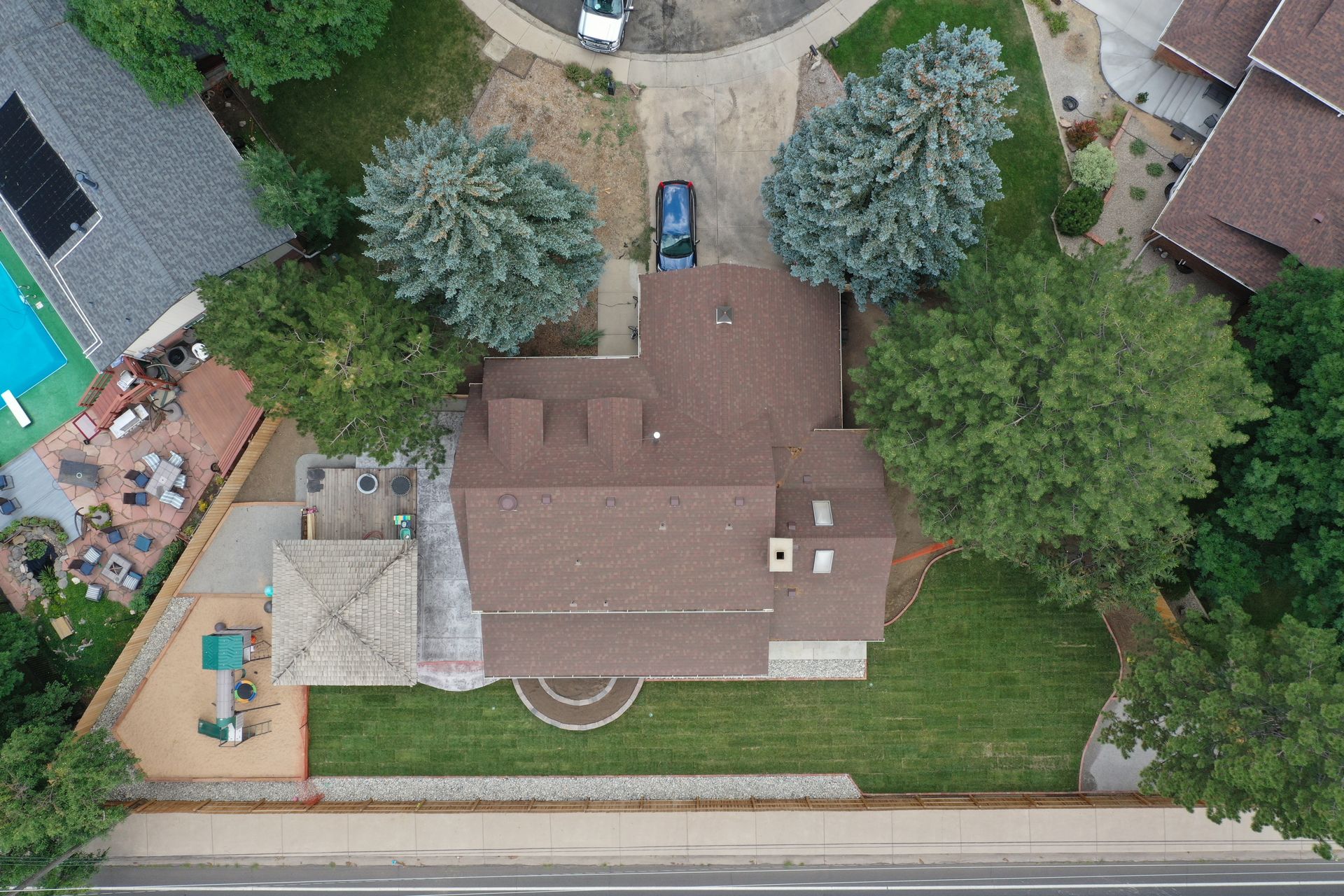 Aerial view of a brown-roofed house with trees, green lawn, driveway, and a backyard patio with a pool.