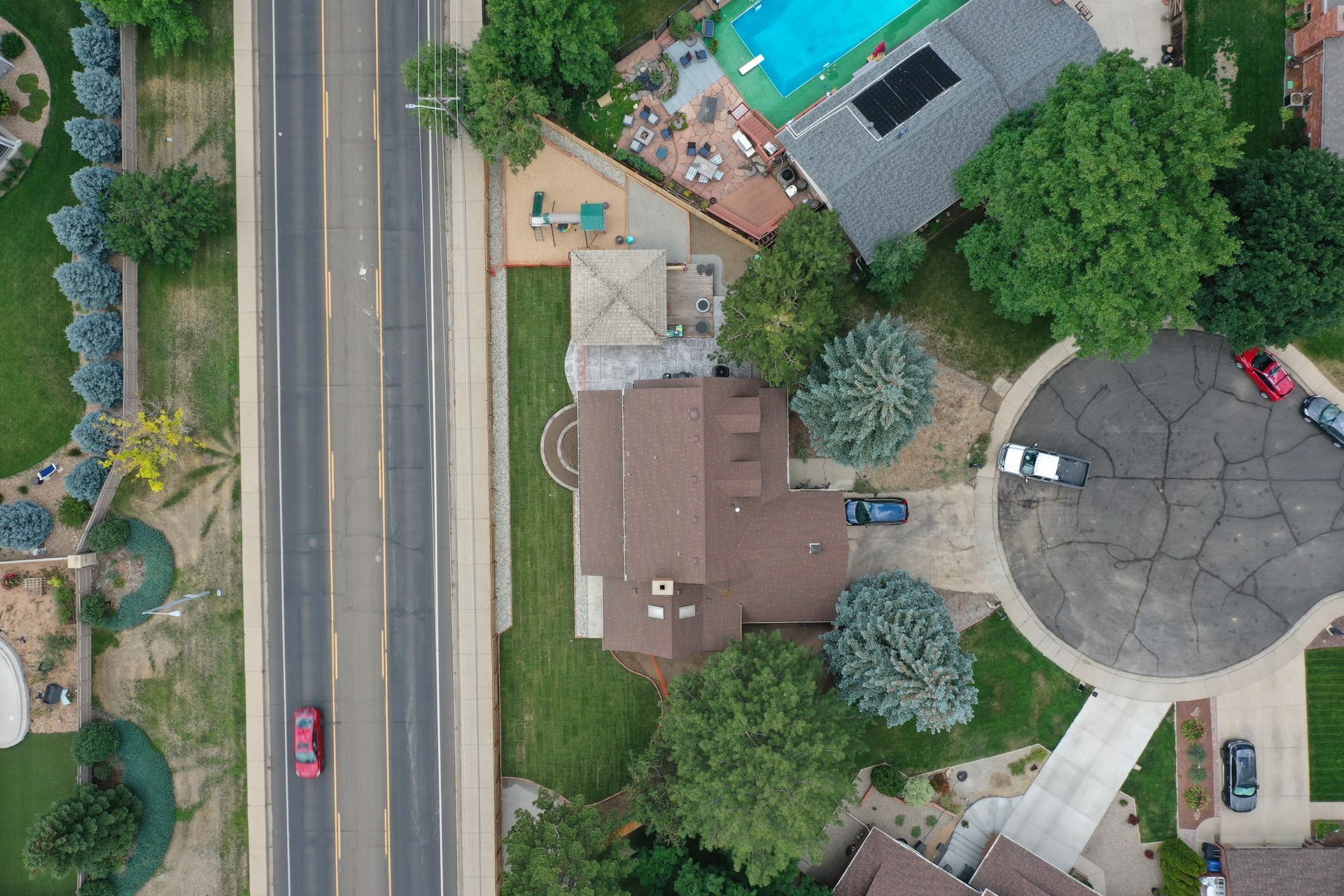 Overhead view of a house next to a road, with cars, trees, a pool, and houses.