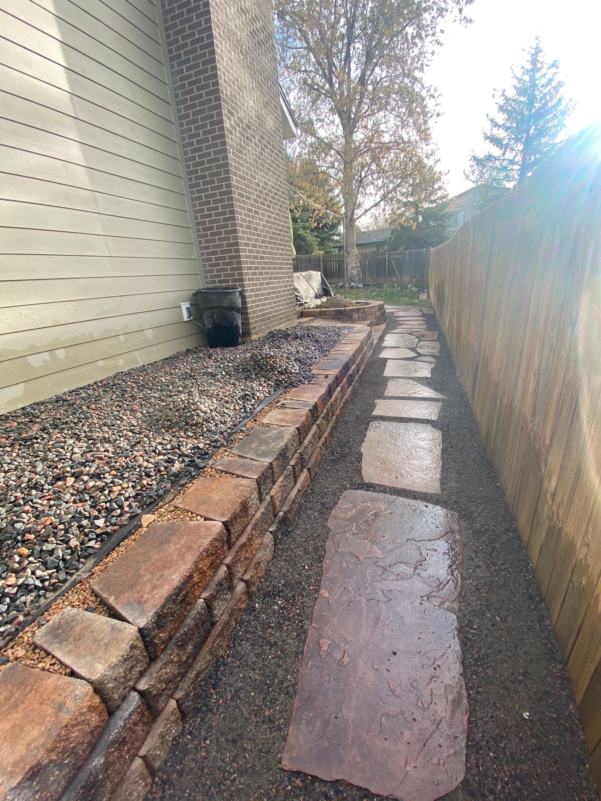 Stone path between a brick retaining wall and a wood fence, alongside a building with siding.