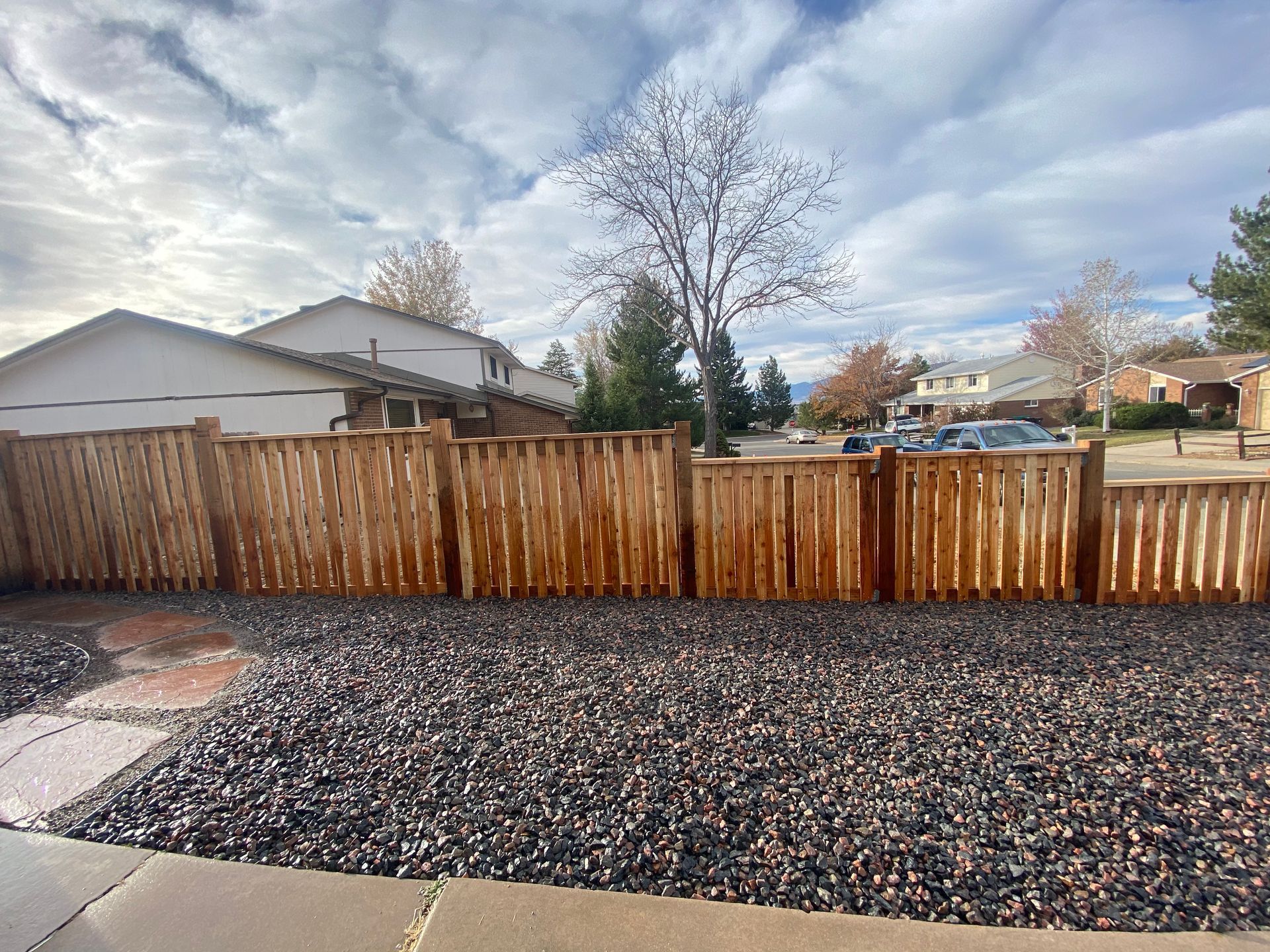 Wooden fence in front of a yard with dark gravel. Houses and a tree are visible in the background.