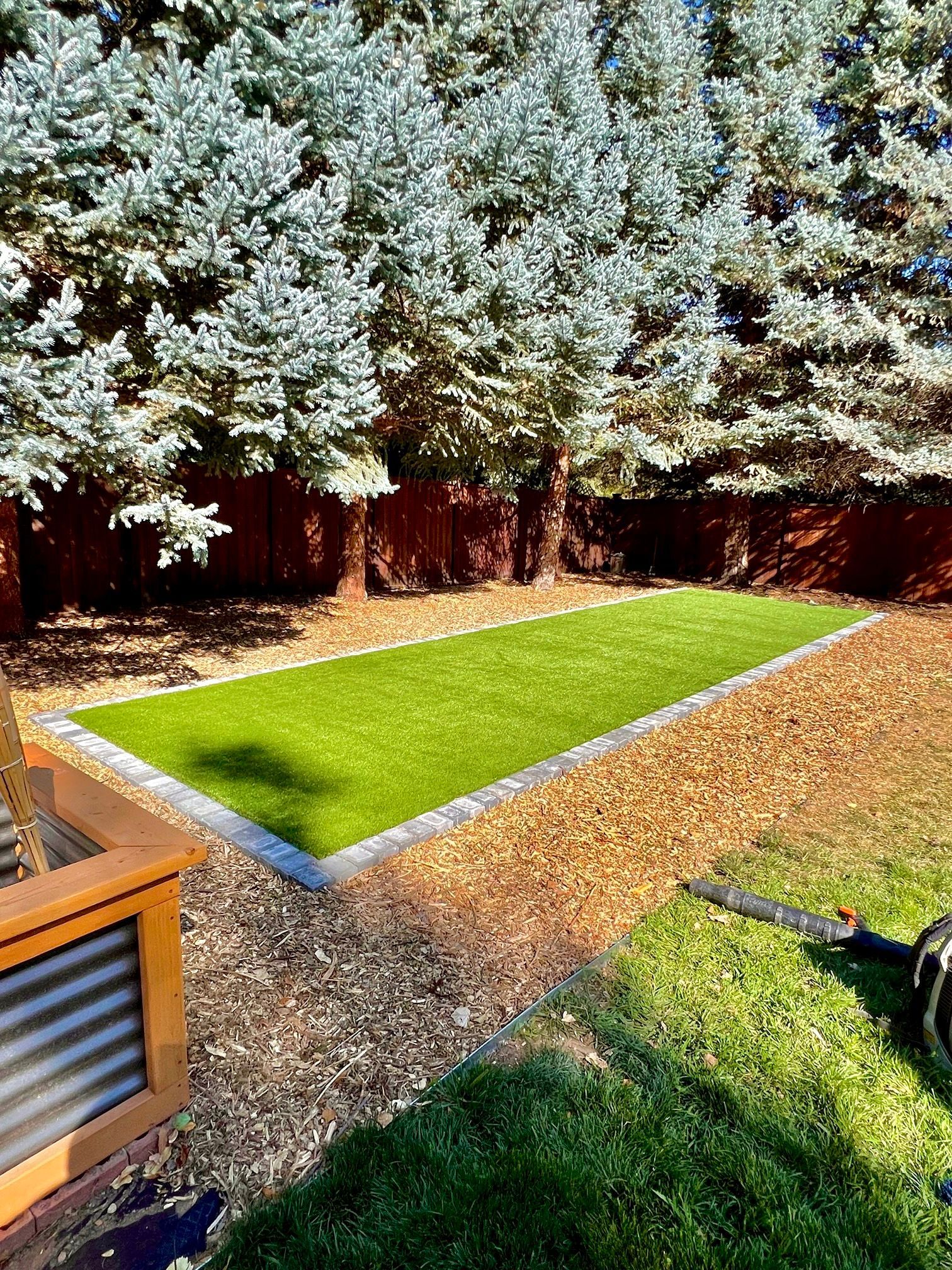 Artificial turf rectangle bordered by concrete and wood chips, framed by evergreen trees.