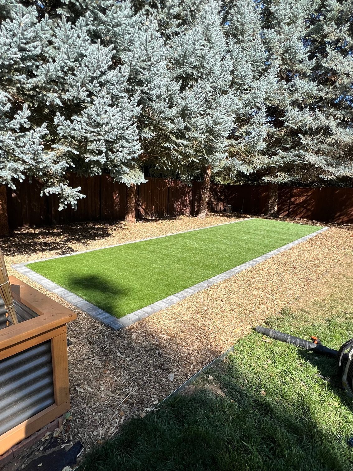 A rectangular artificial turf area surrounded by wood chips, trees in background, brown fence.