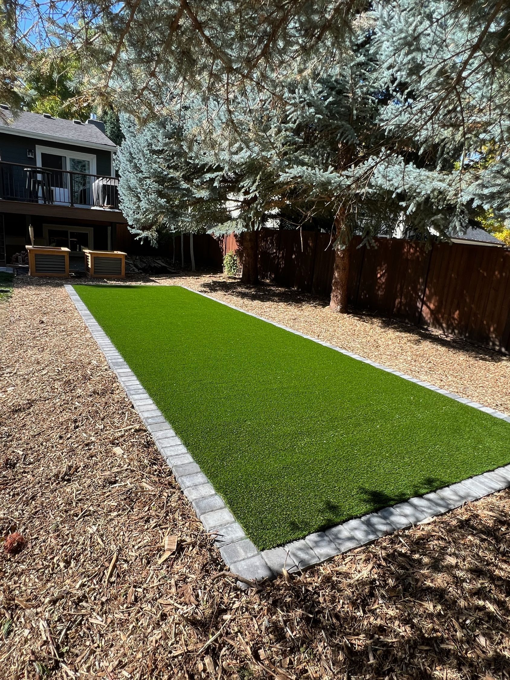 Rectangular artificial turf area bordered by pavers in a backyard with a deck and trees.
