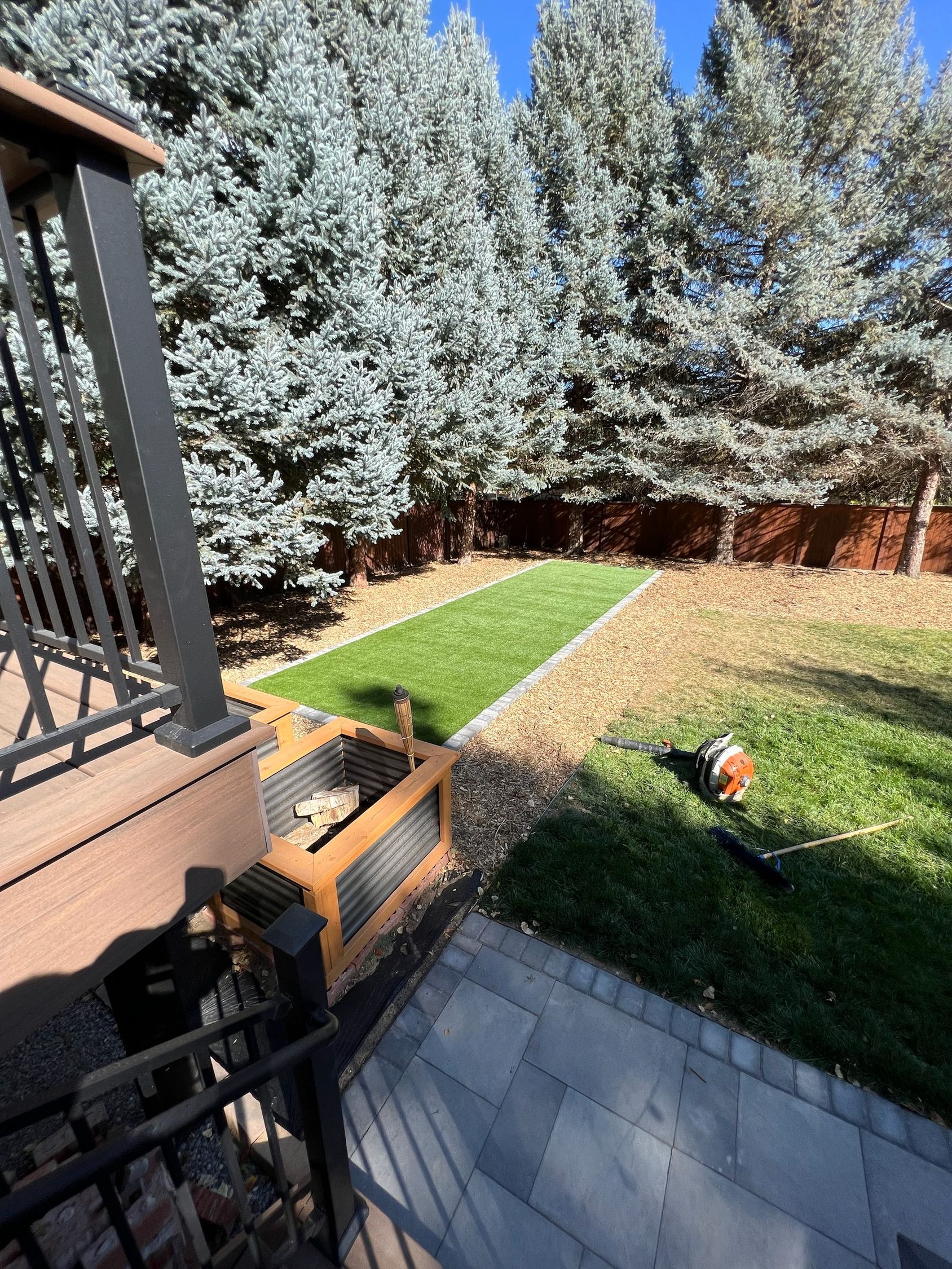 Backyard with green artificial turf strip bordered by rocks, leading to trees; a patio and deck are in view.