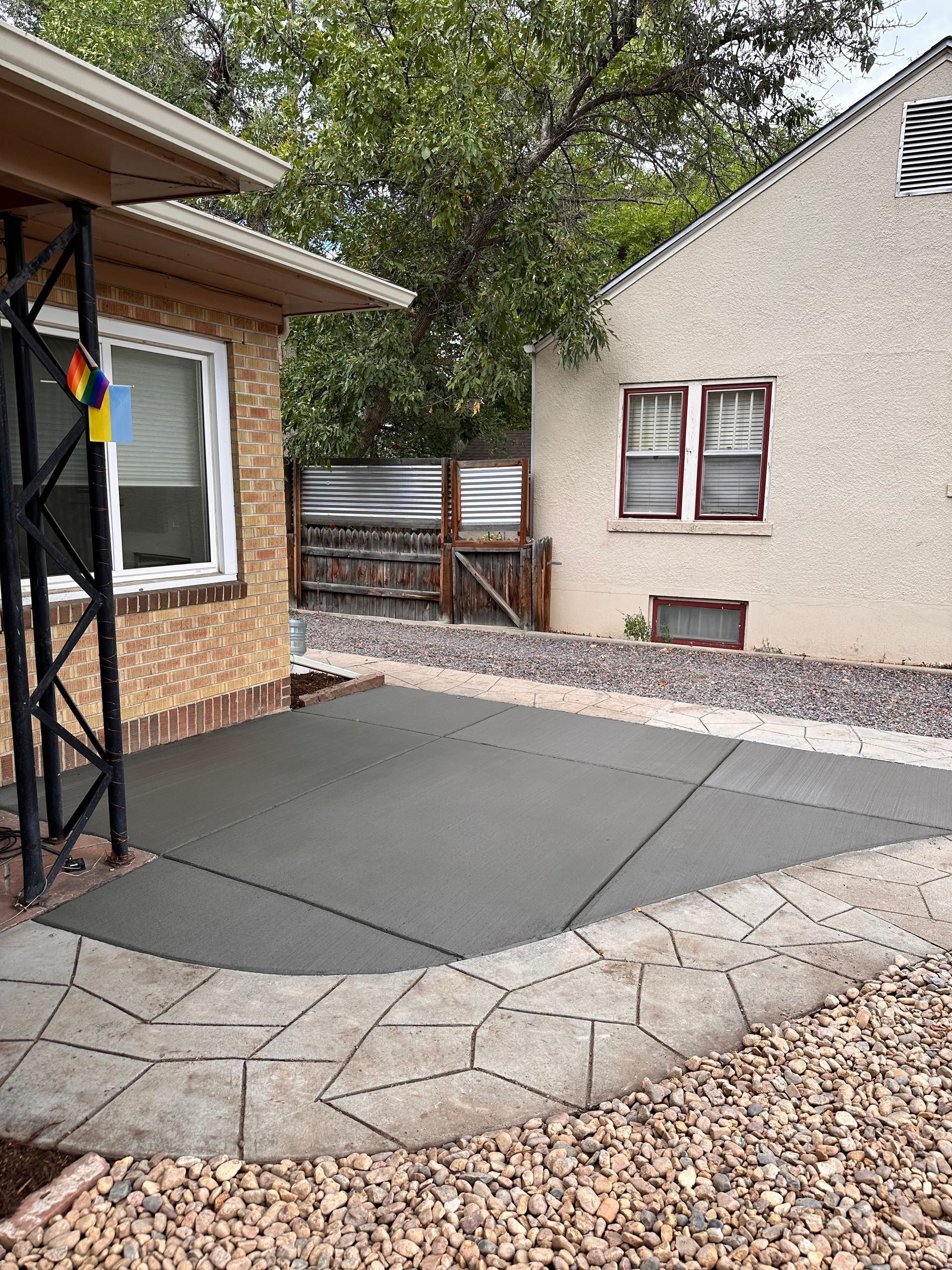 Concrete patio next to a brick house and a stucco building, bordered by stepping stones and gravel.