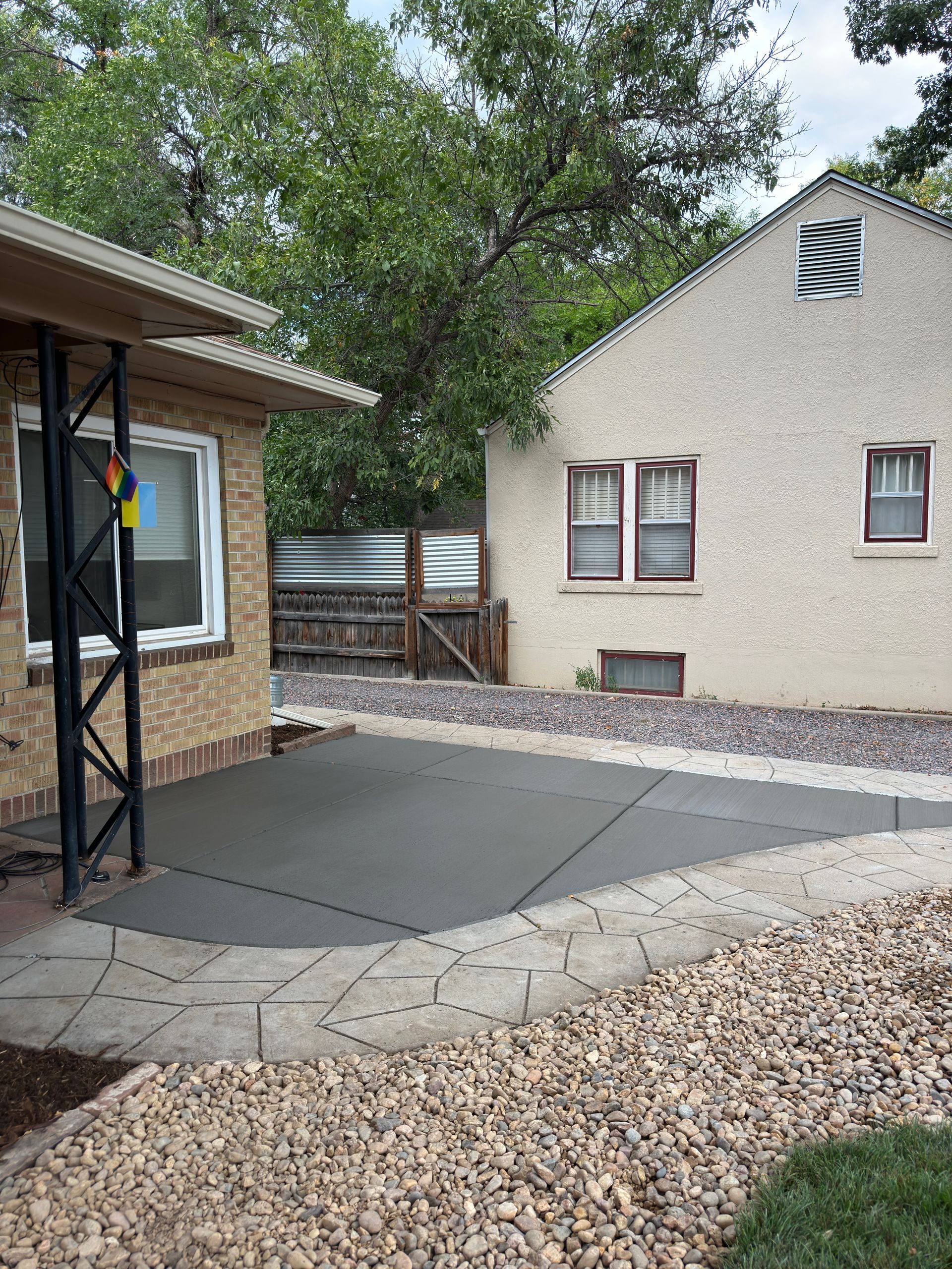 Freshly poured concrete patio next to a house and detached garage. Gravel border with trees in background.
