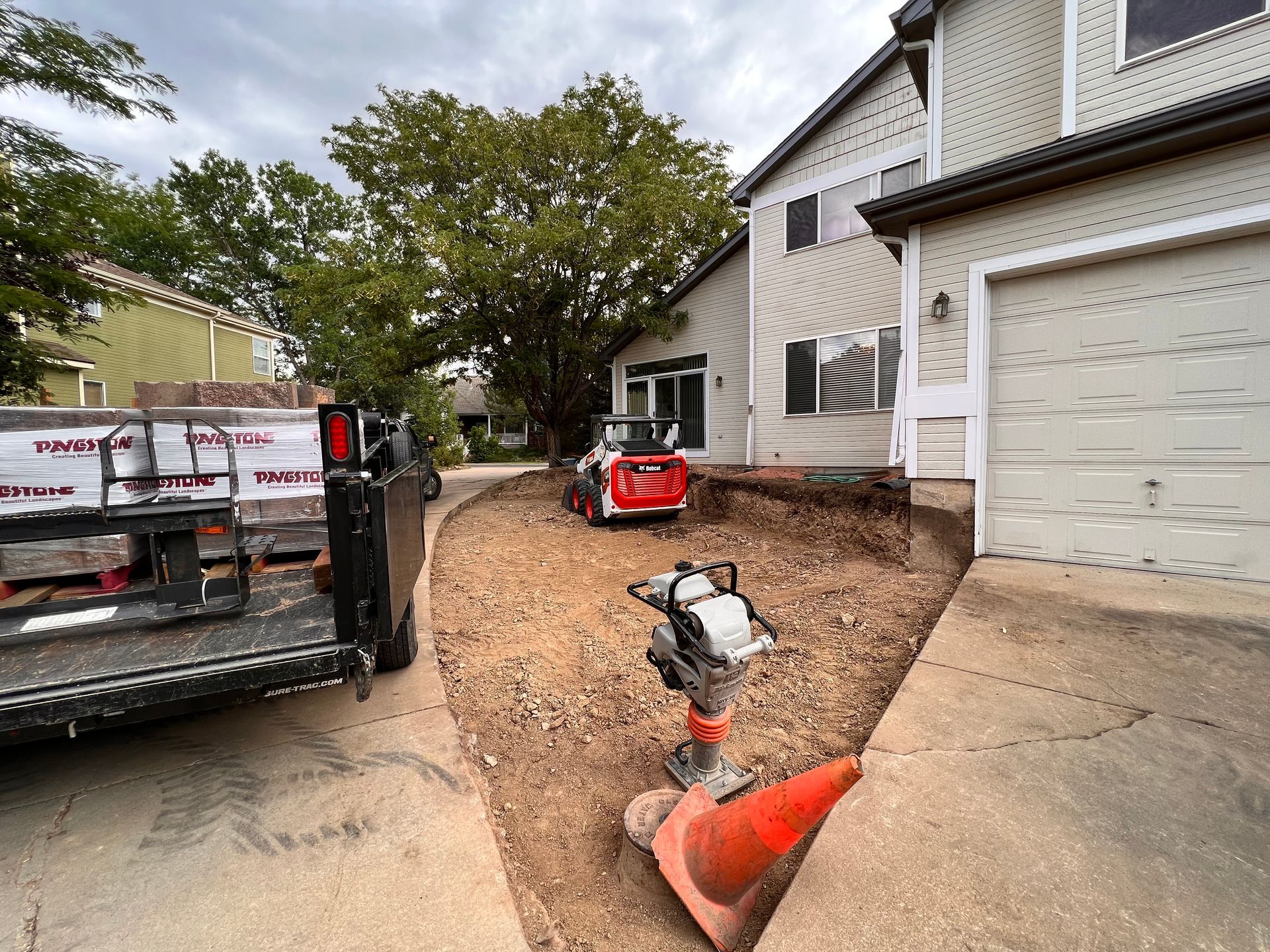 Construction site with a mini excavator, trailer, and compactor near a house.