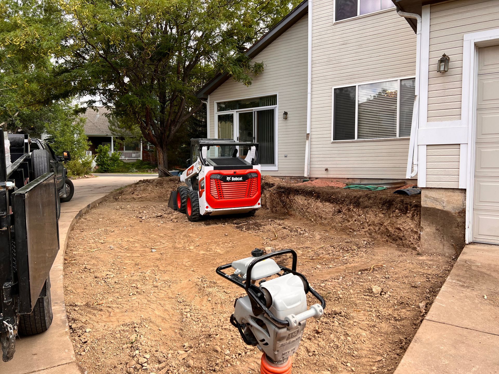 Bobcat skid-steer and plate compactor on construction site near a two-story house with dirt and gravel.