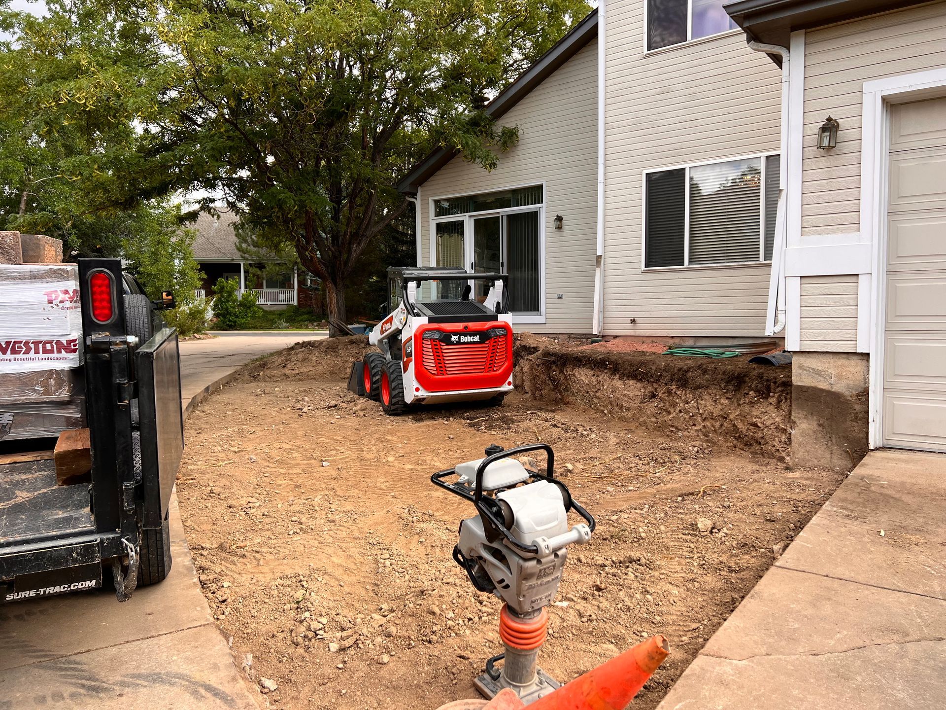 A Bobcat and compactor working on a dirt area near a house and garage; a truck is parked nearby.