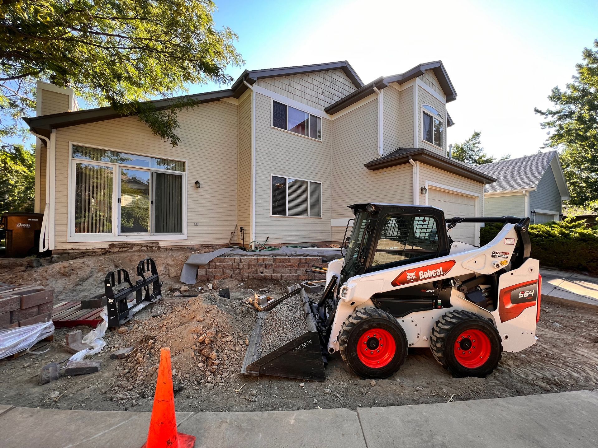 Bobcat skid-steer loader on construction site in front of a house, soil and brick visible, cone for safety.