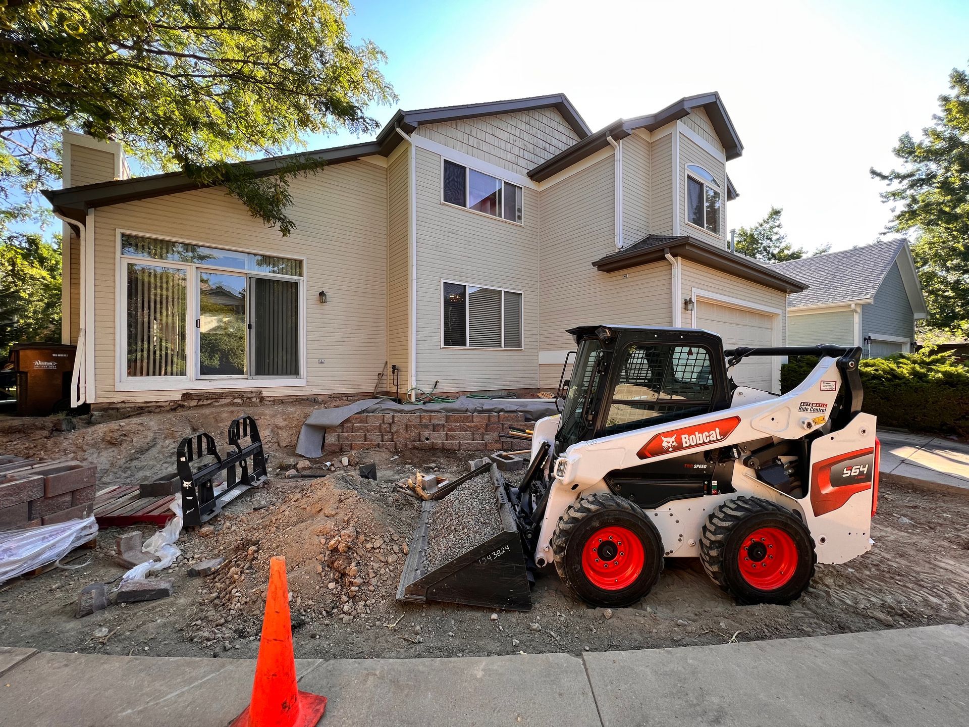 Bobcat skid-steer loader working near a two-story beige house during construction with an orange traffic cone in front.