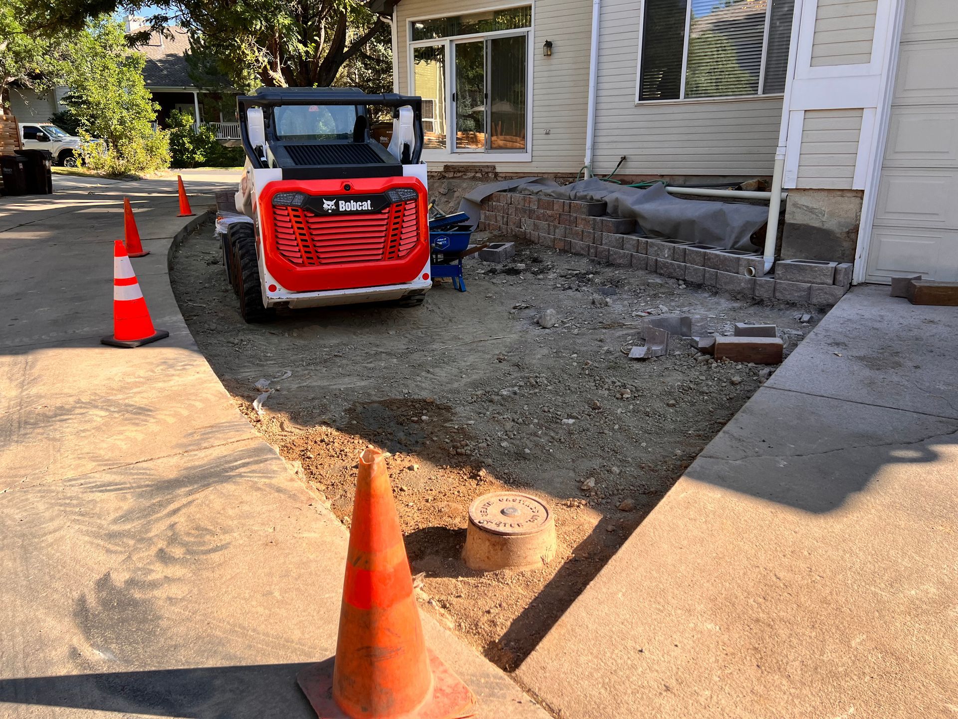 Bobcat on gravel near house, orange cones mark pathway.