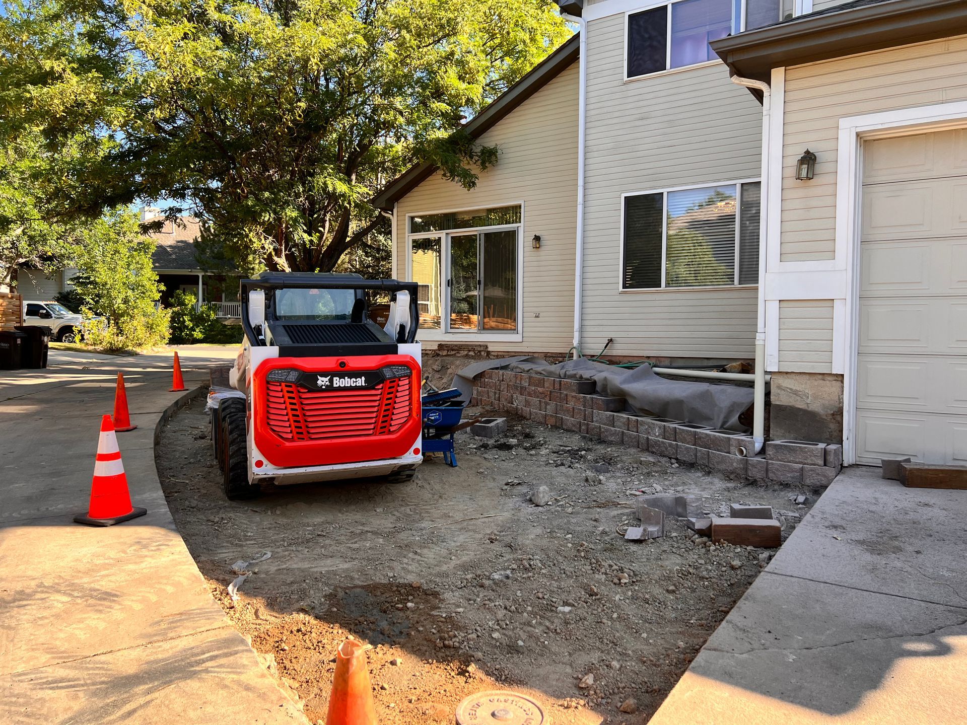 A Bobcat skid-steer loader working near a house, with construction materials and orange cones visible.