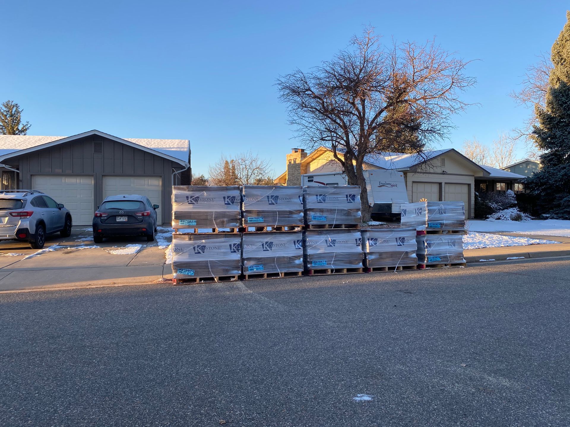 Pallets of building materials on a snow-covered street in front of houses on a sunny day.