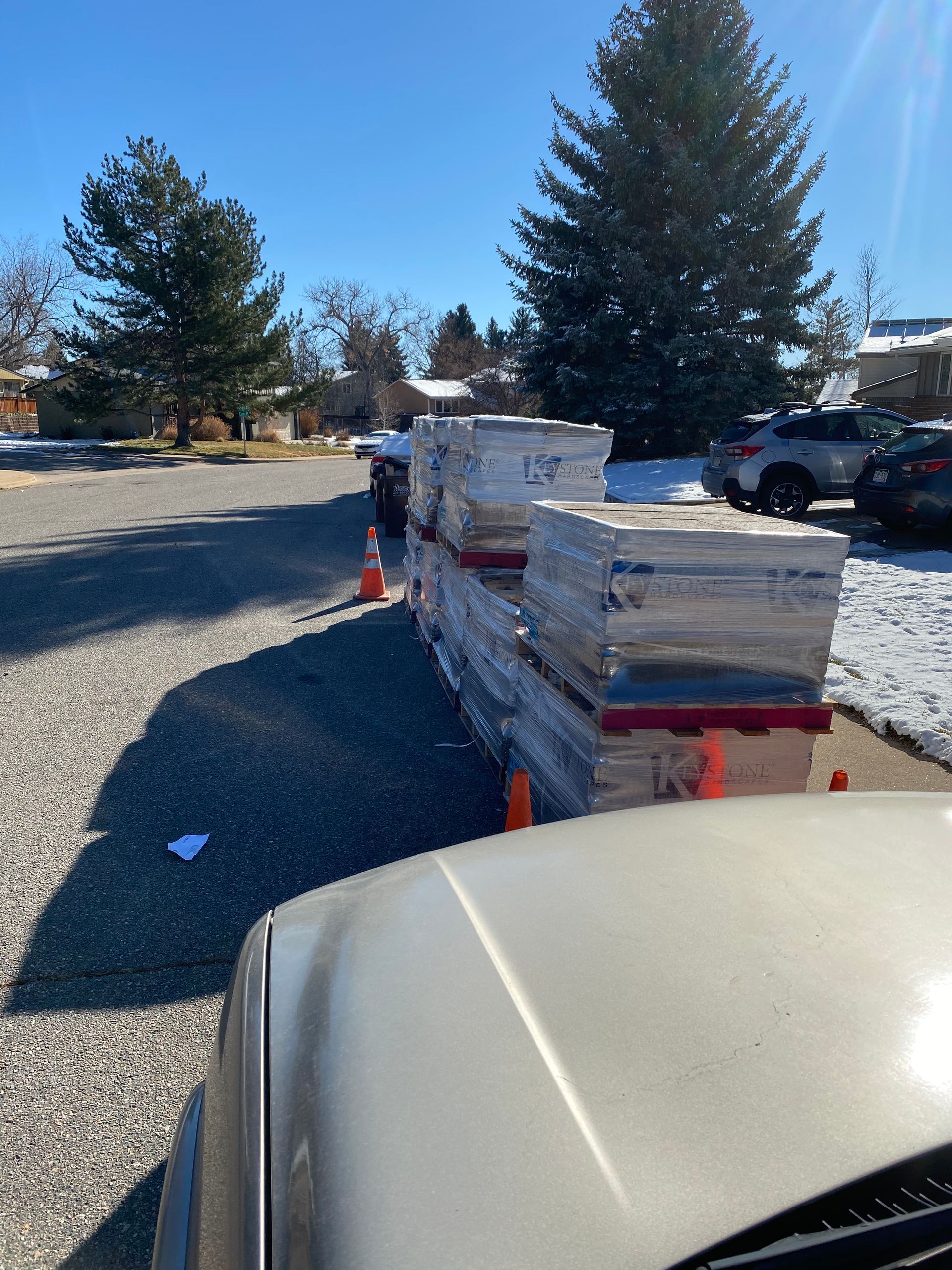 Stacks of wrapped building materials on a gravel driveway, orange cones, parked cars, sunny day, snowy ground.