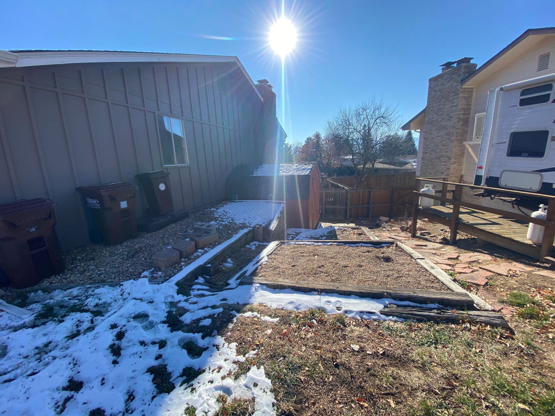 Backyard with snow, brown buildings, wooden fence, sun shining, trash cans.