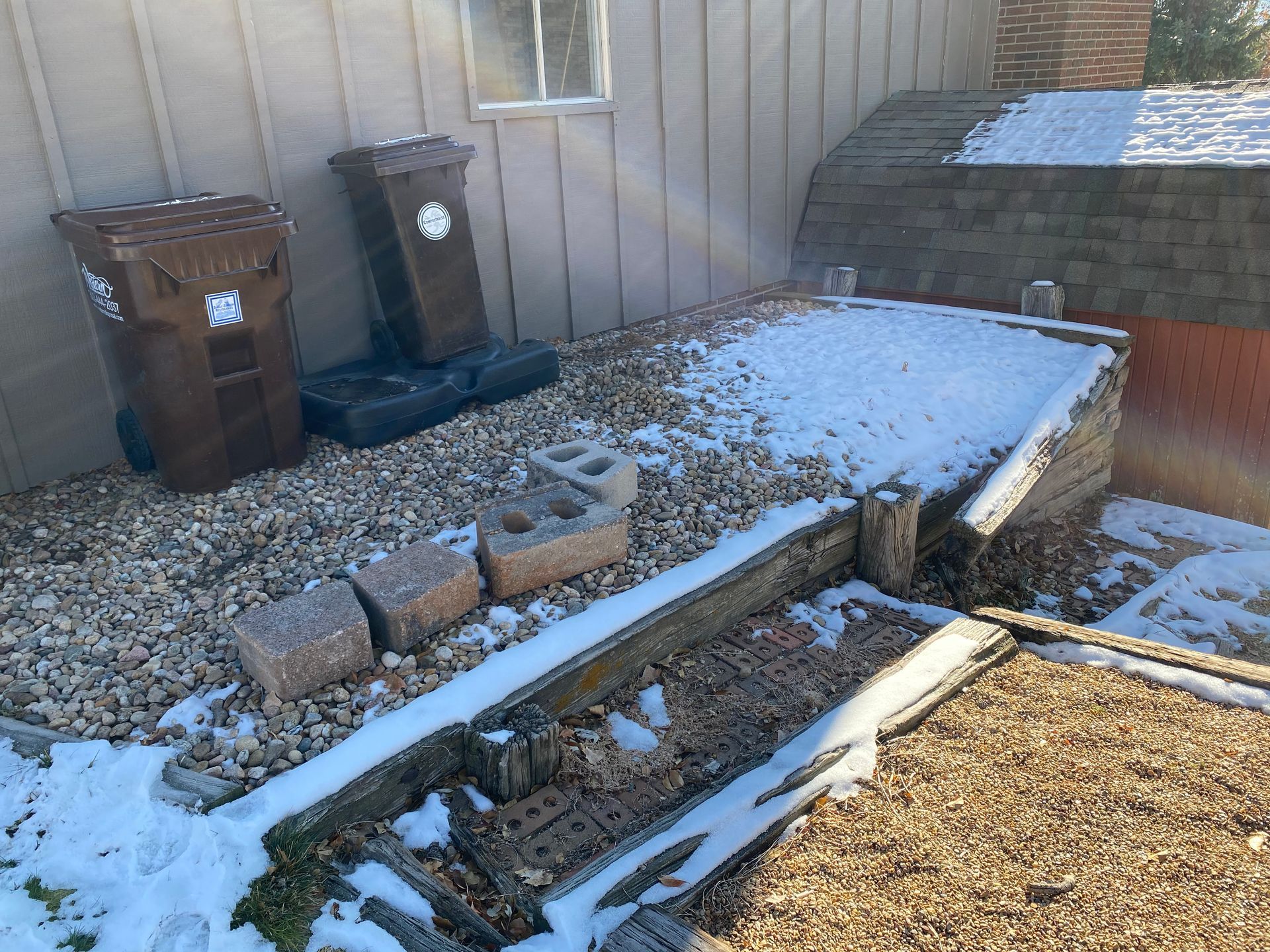 Brown trash cans next to a snow-covered wooden structure and gravel ground. Sunny, outdoor setting.