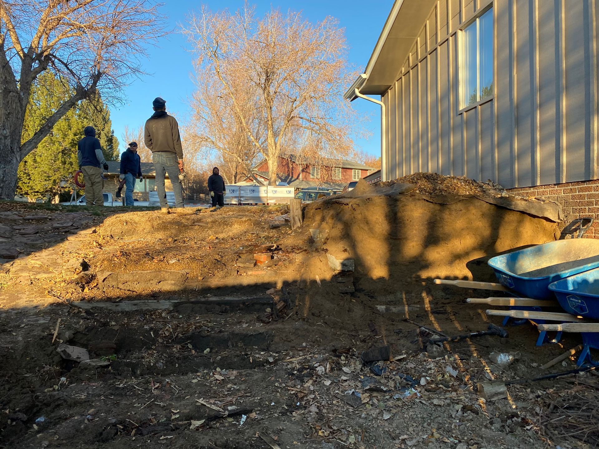 People working on a landscaping project near a house; dirt pile and wheelbarrows are visible.