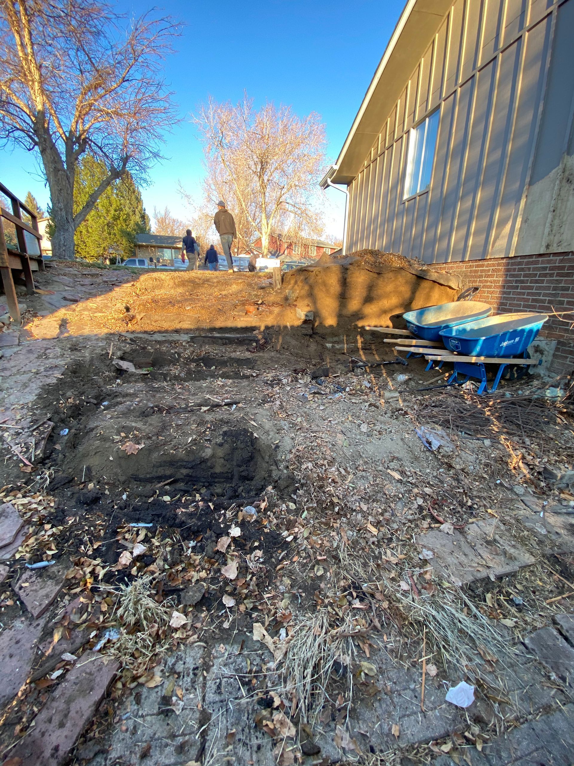 Backyard excavation: dirt pile, exposed ground, blue wheelbarrow, people in the distance, sunny day.