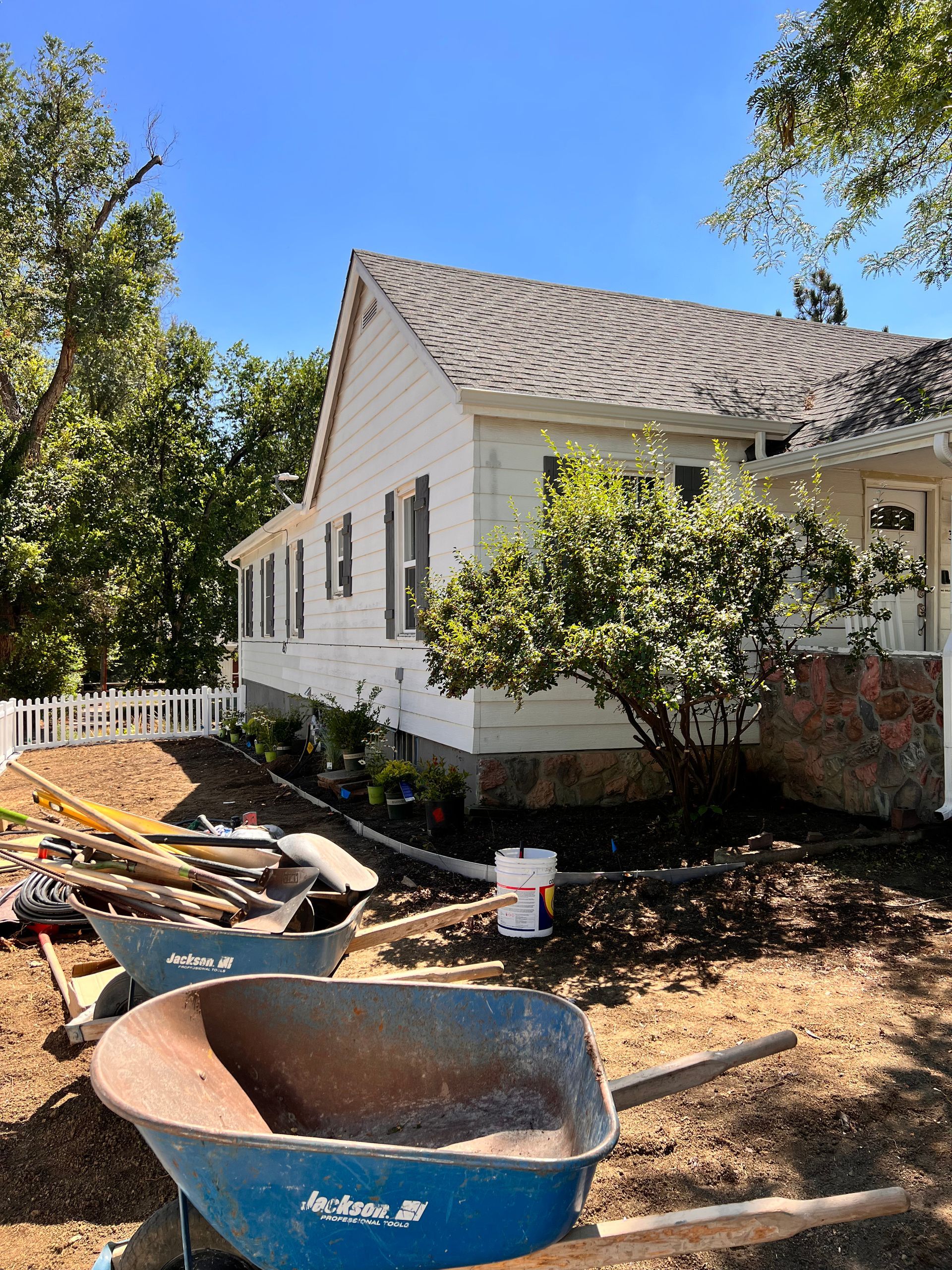 Exterior view of a white house under construction with wheelbarrows and tools in the yard on a sunny day.