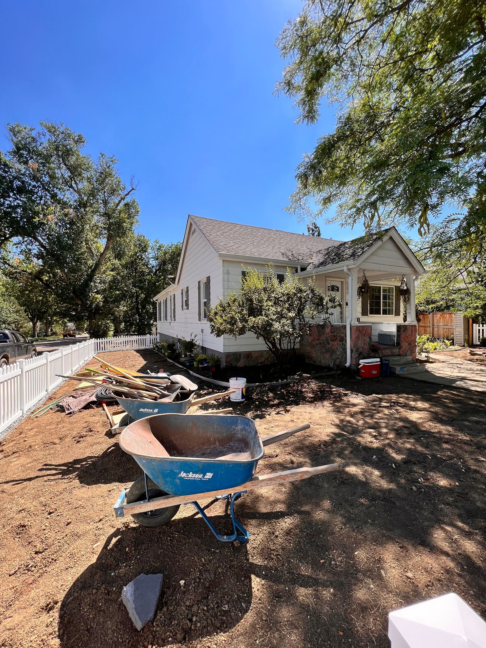 House under construction, dirt yard, blue wheelbarrow, white fence, trees, sunny day.