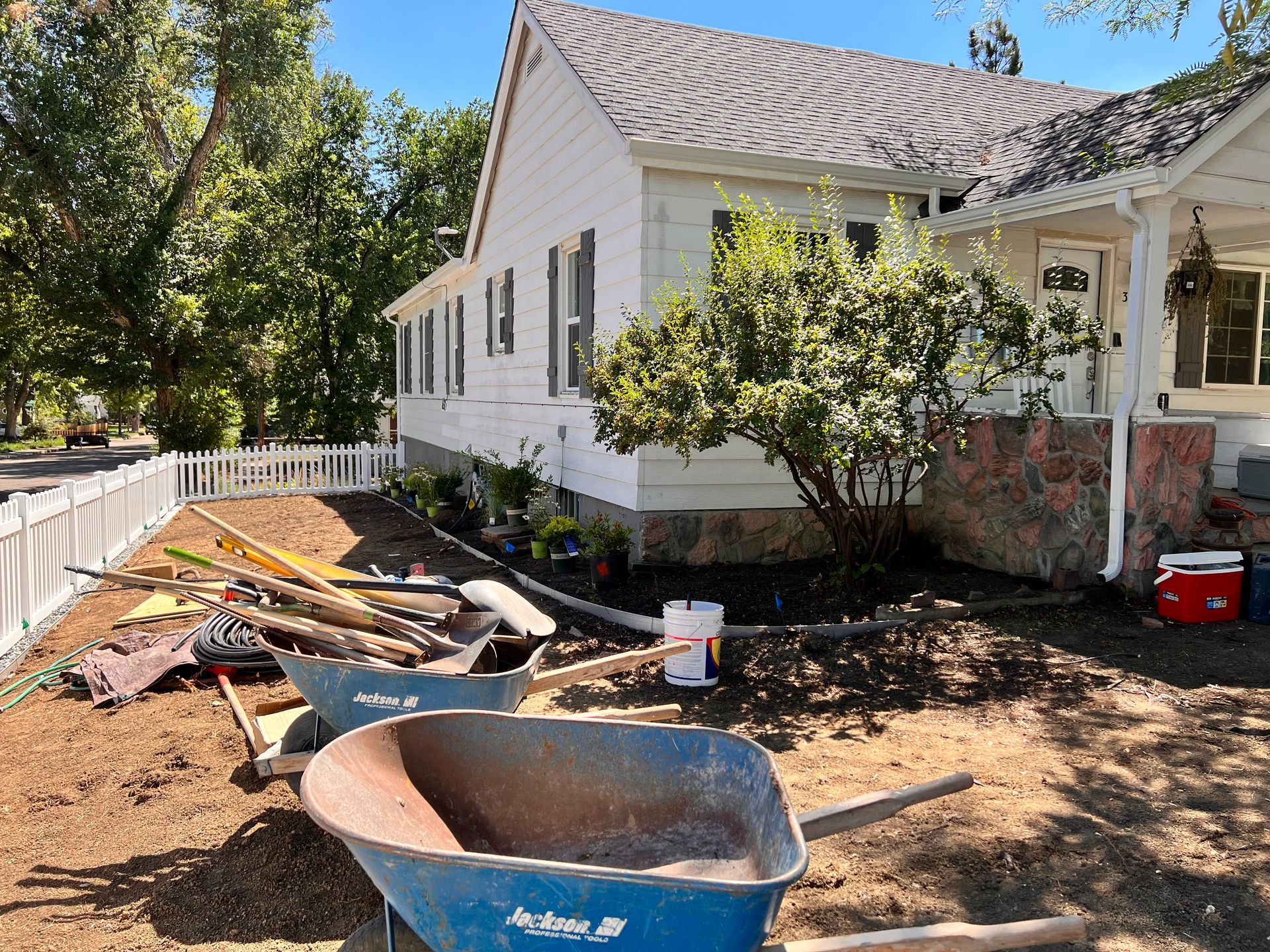 House with landscaping under construction; wheelbarrows, tools, dirt, and plants in yard.