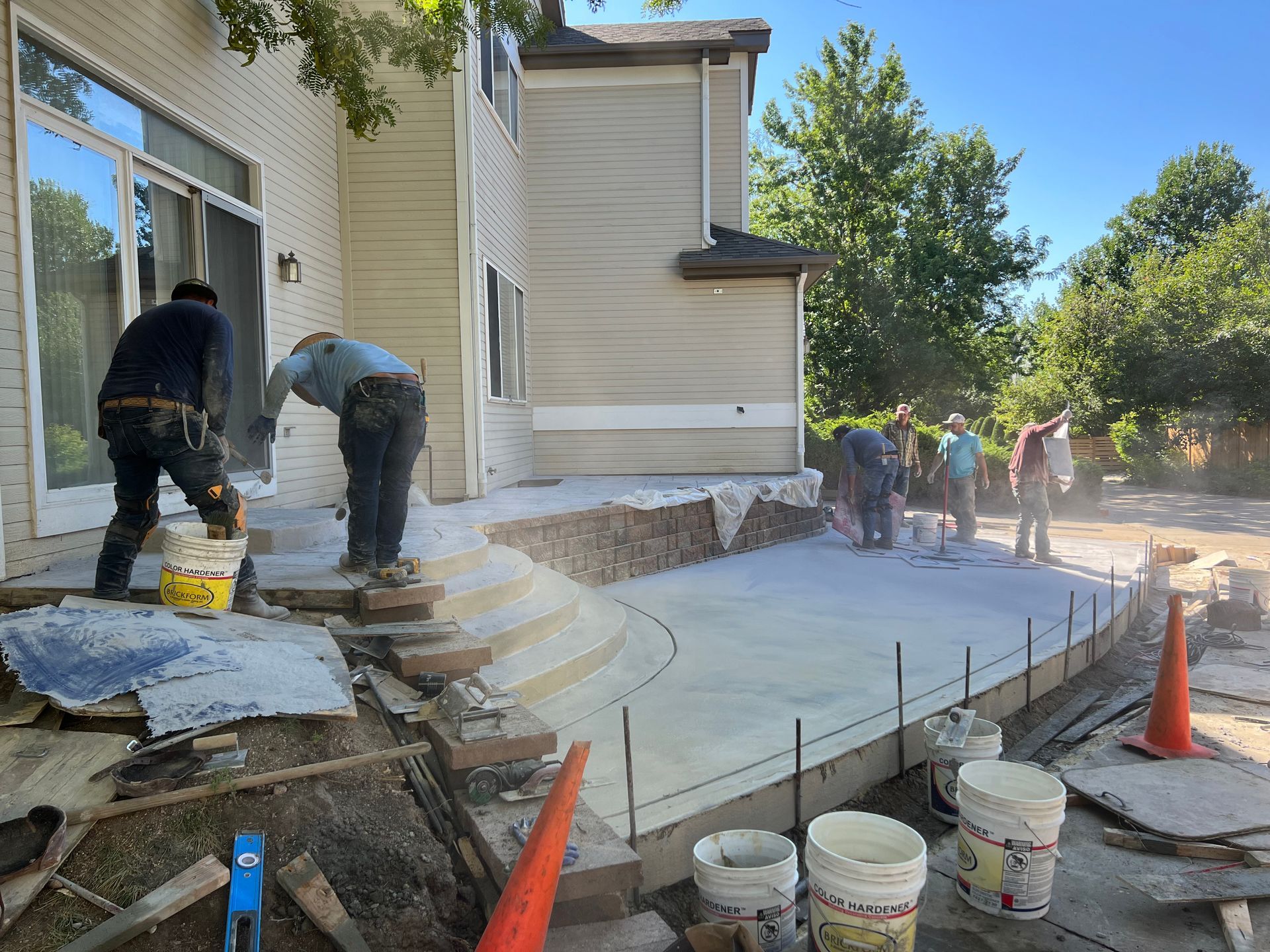 Construction workers pouring concrete for a patio next to a two-story beige house; sunny day.