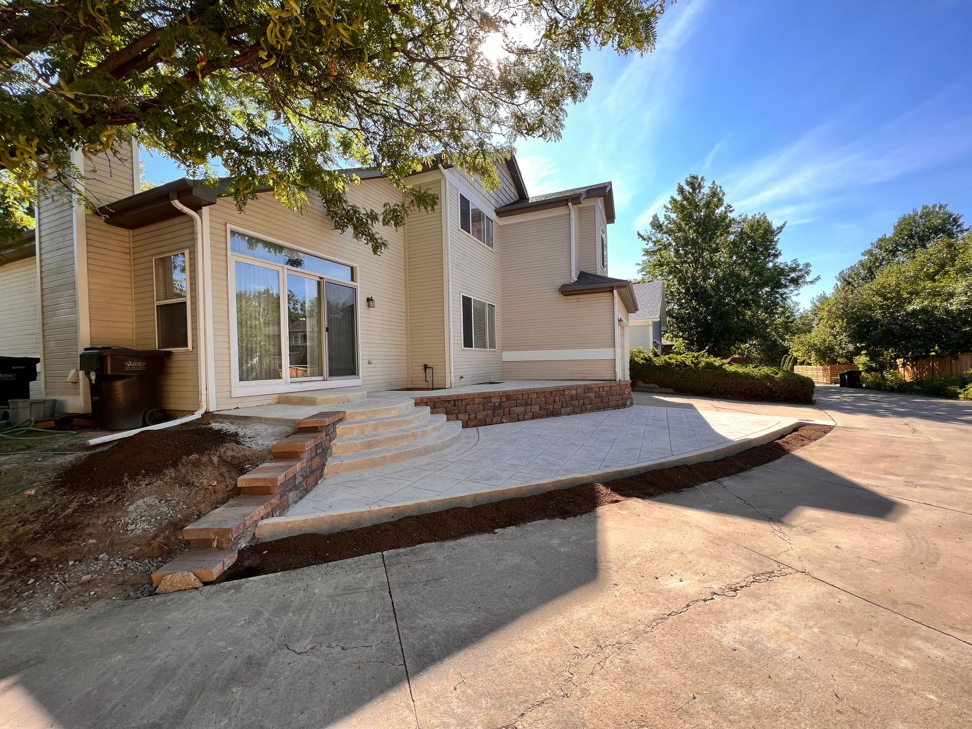 House exterior with stone patio, steps, and driveway. Sunny day with trees and blue sky.