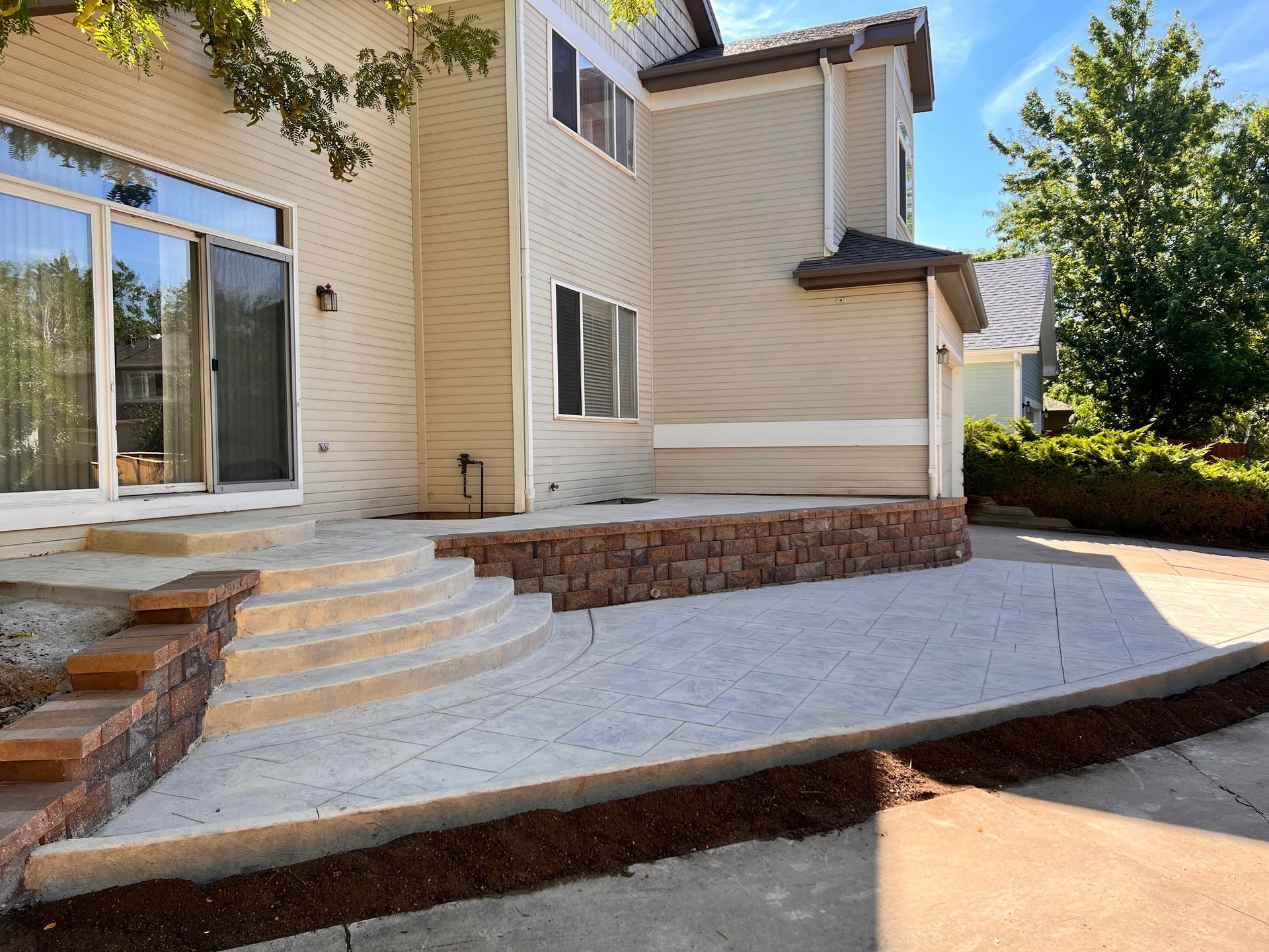 A two-story beige house with a concrete patio, retaining wall, and steps.