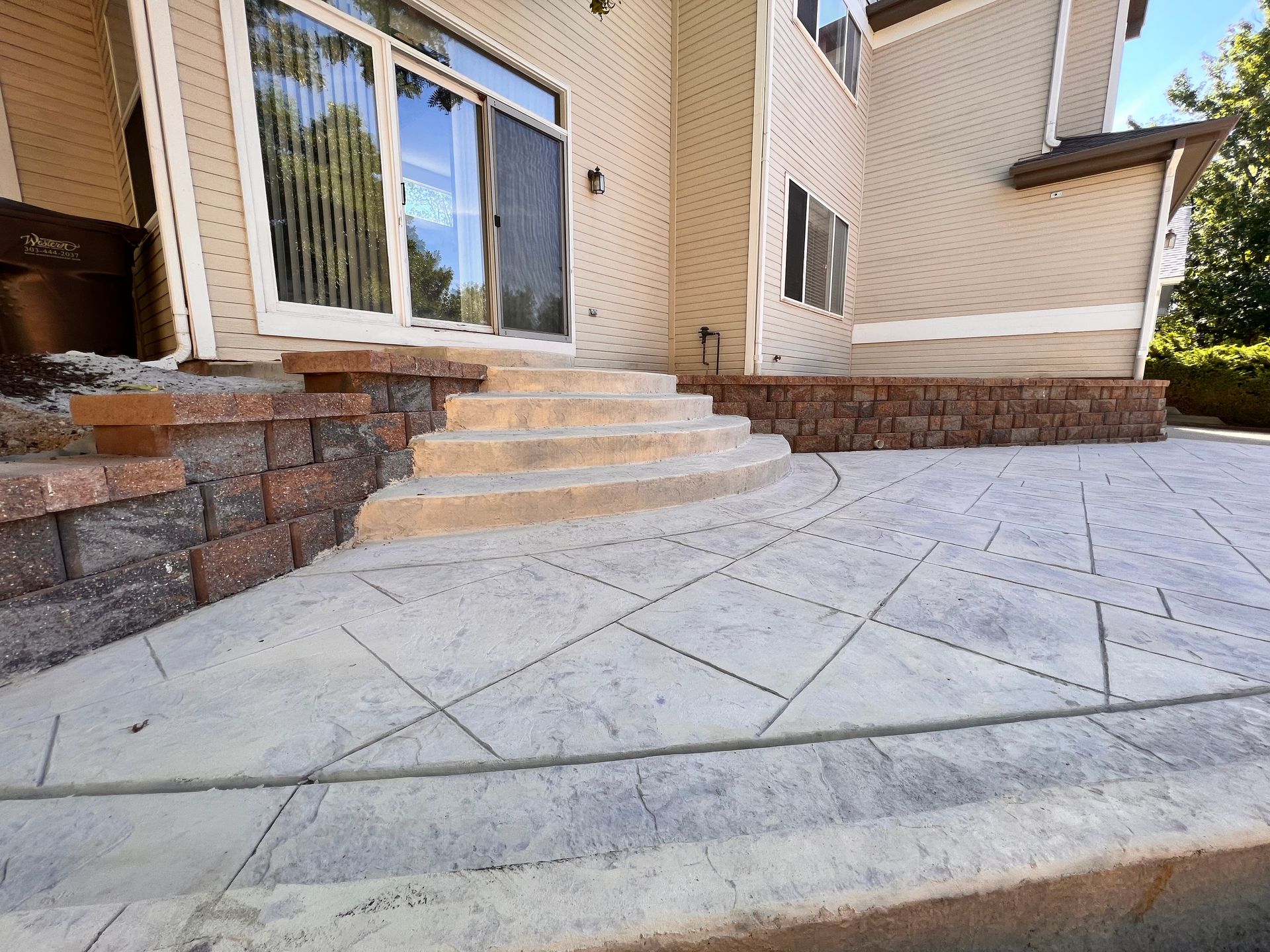 Outdoor concrete patio with steps and retaining walls; tan house in the background.