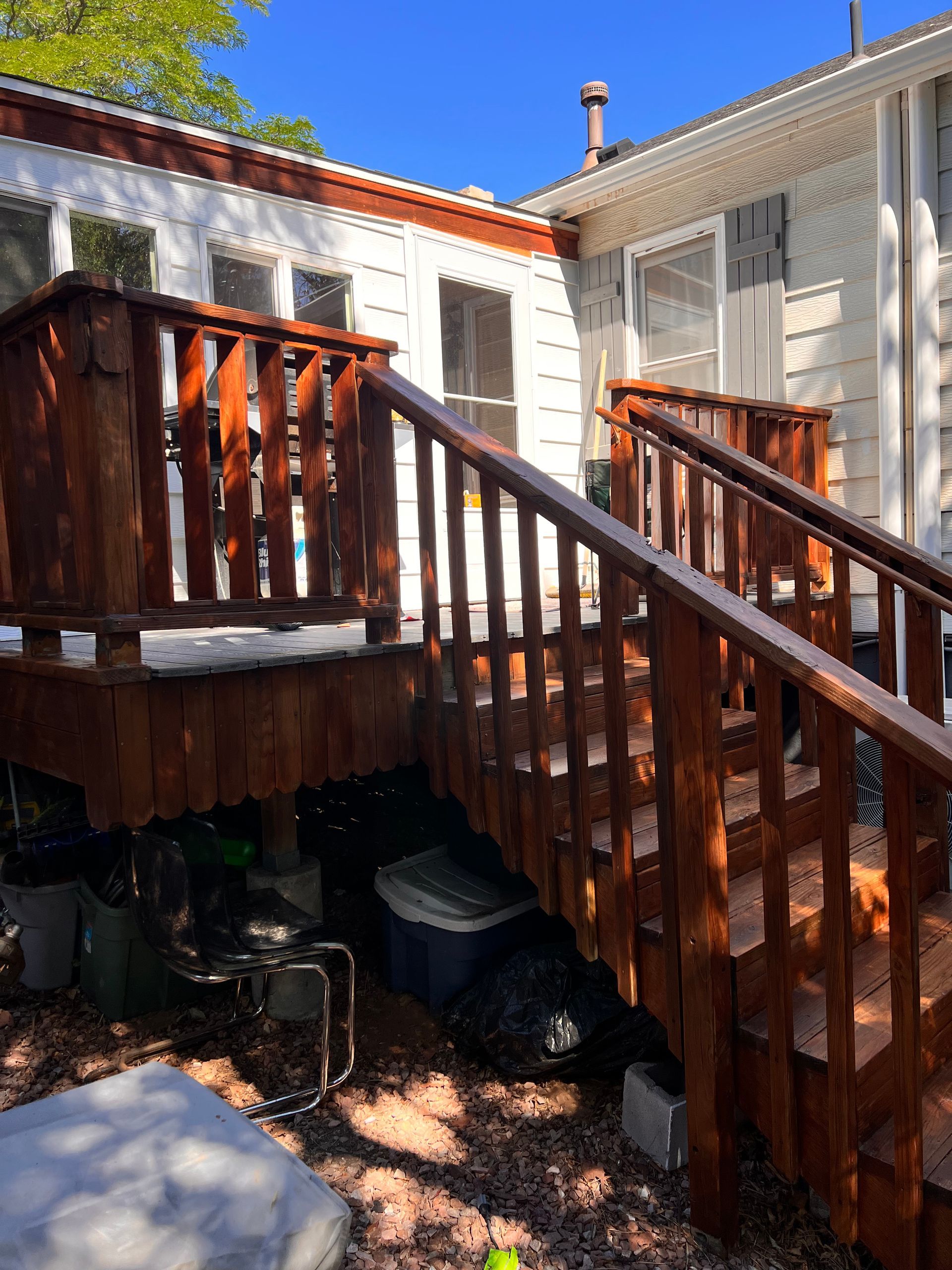 Wooden deck with stairs, stained reddish-brown, leading up to a white house with windows. Sunny day.