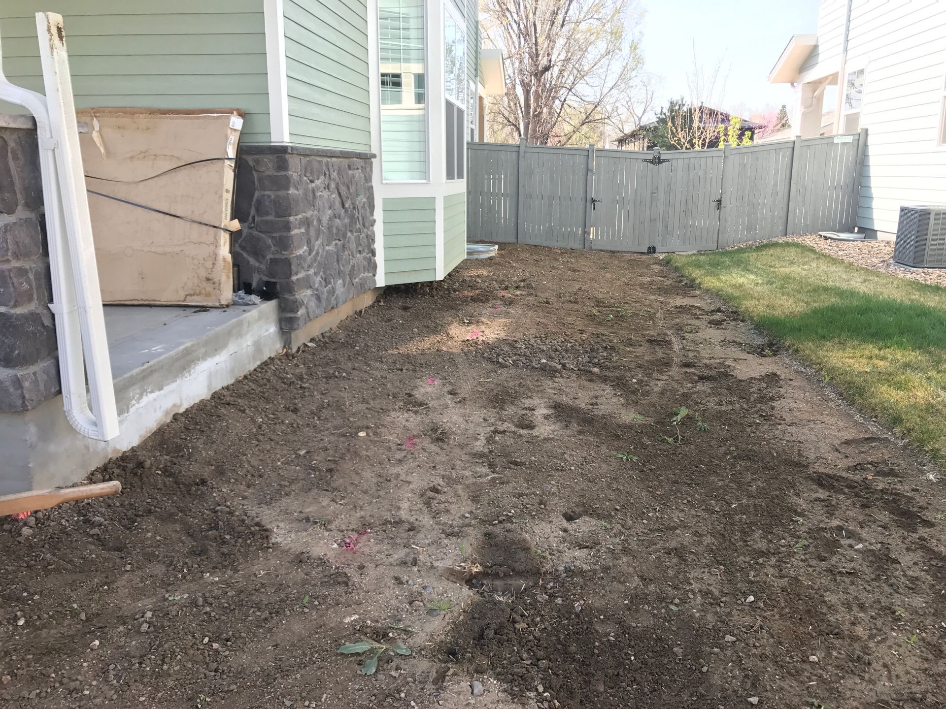 A front yard with dirt, next to a house with light green siding and a gray fence.